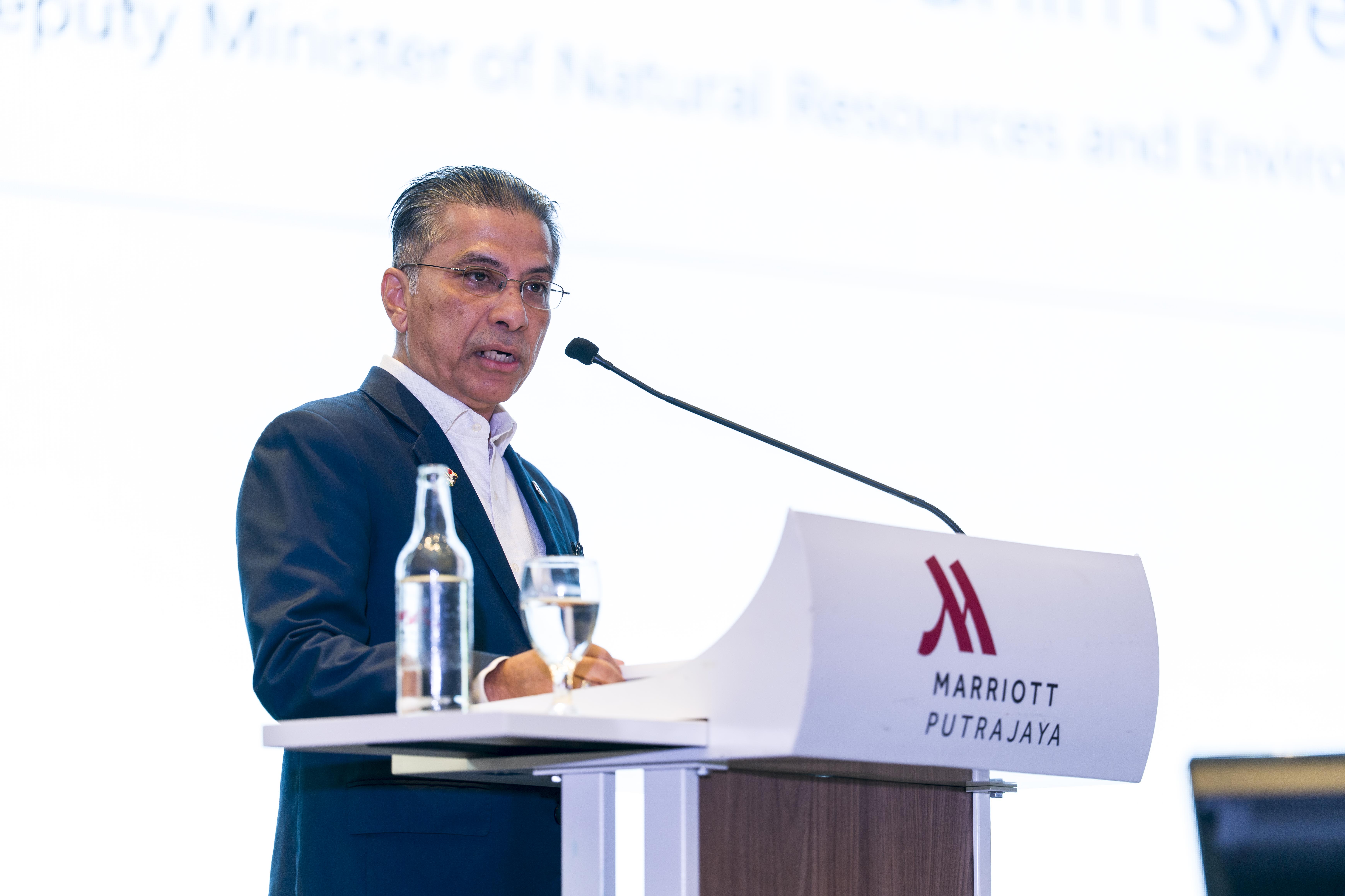 Speaker at podium with microphone and water bottles; blurred face; Marriott logo on lectern.