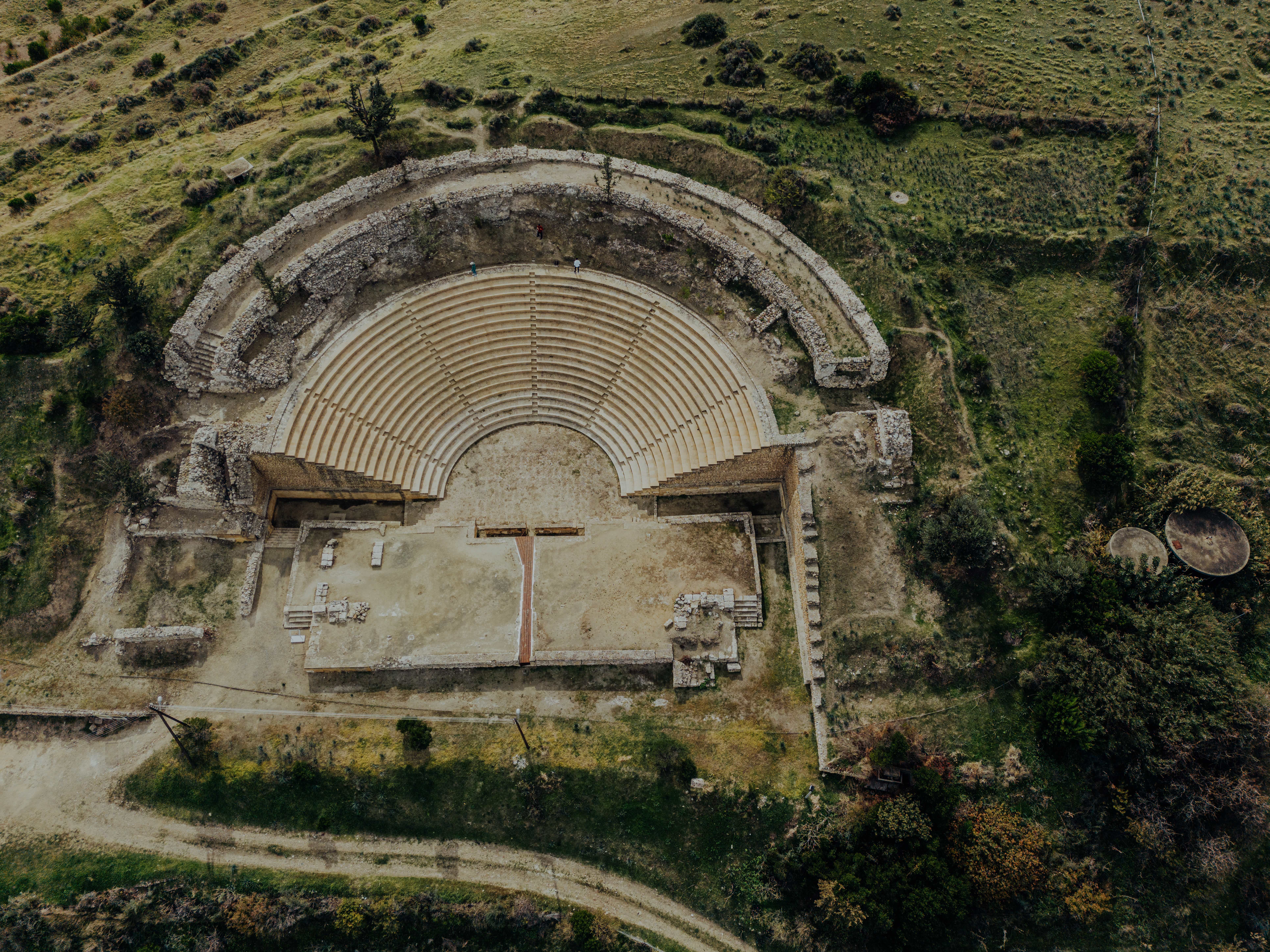 Aerial view of a circular stone amphitheater ruin with tiered seating amid greenery.