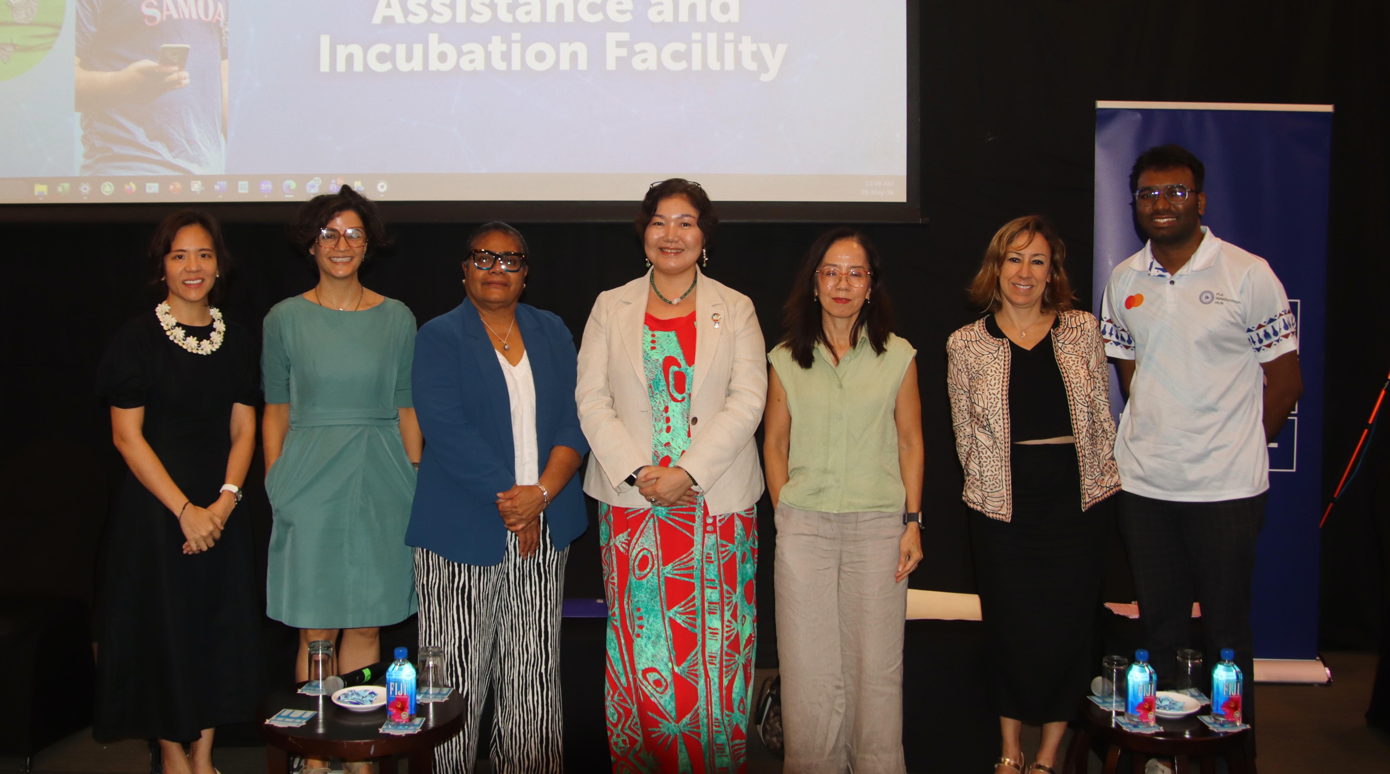 Eight people on a stage in front of a banner reading Assistance and Incubation Facility.