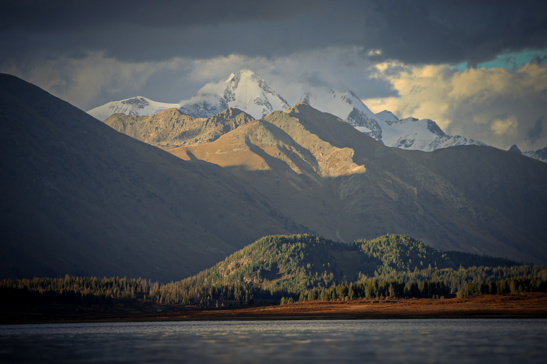 Snow-capped mountains rise over a forested hillside with a lake in the foreground.