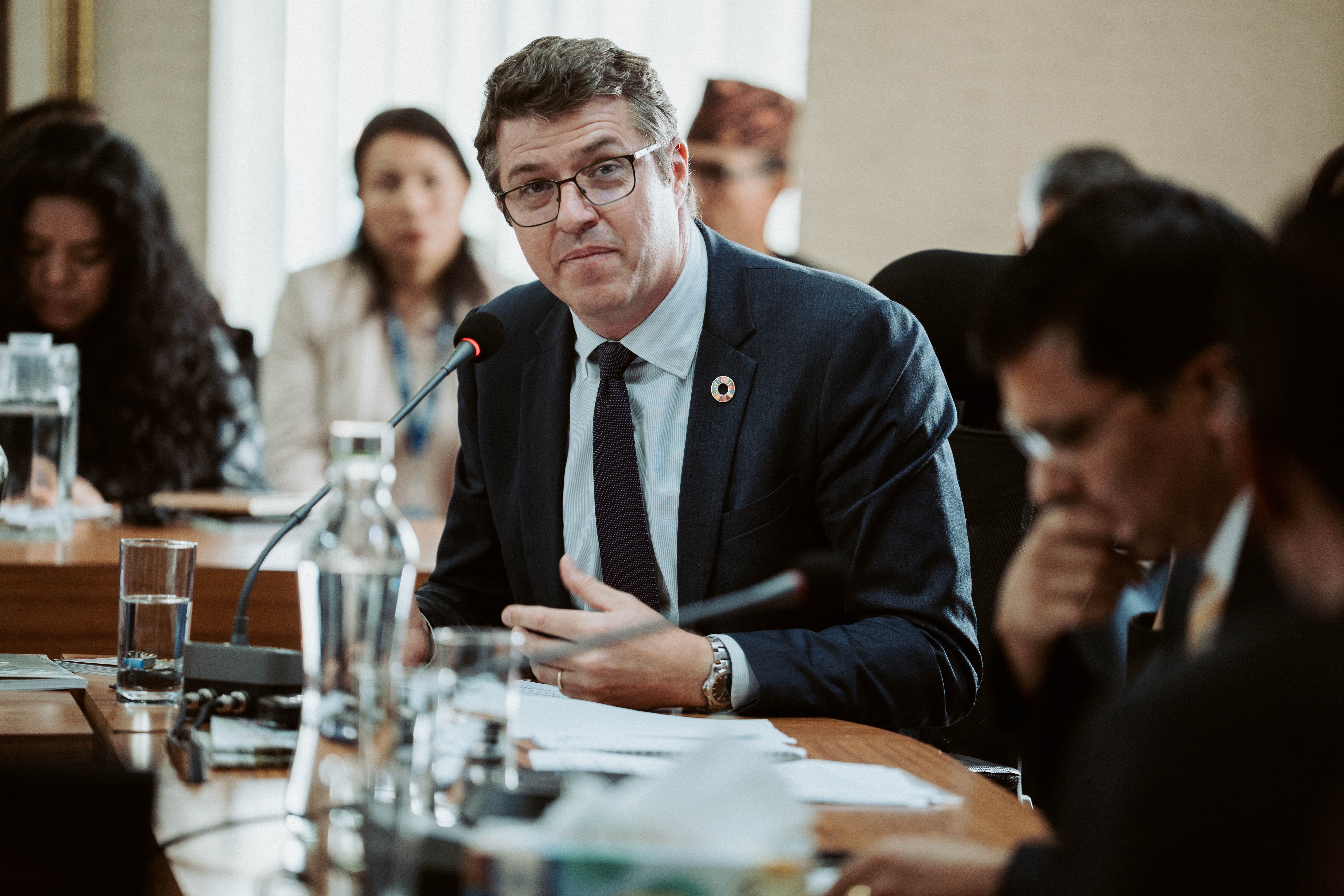 Business meeting around conference table; man in suit speaking, others listening, faces blurred.
