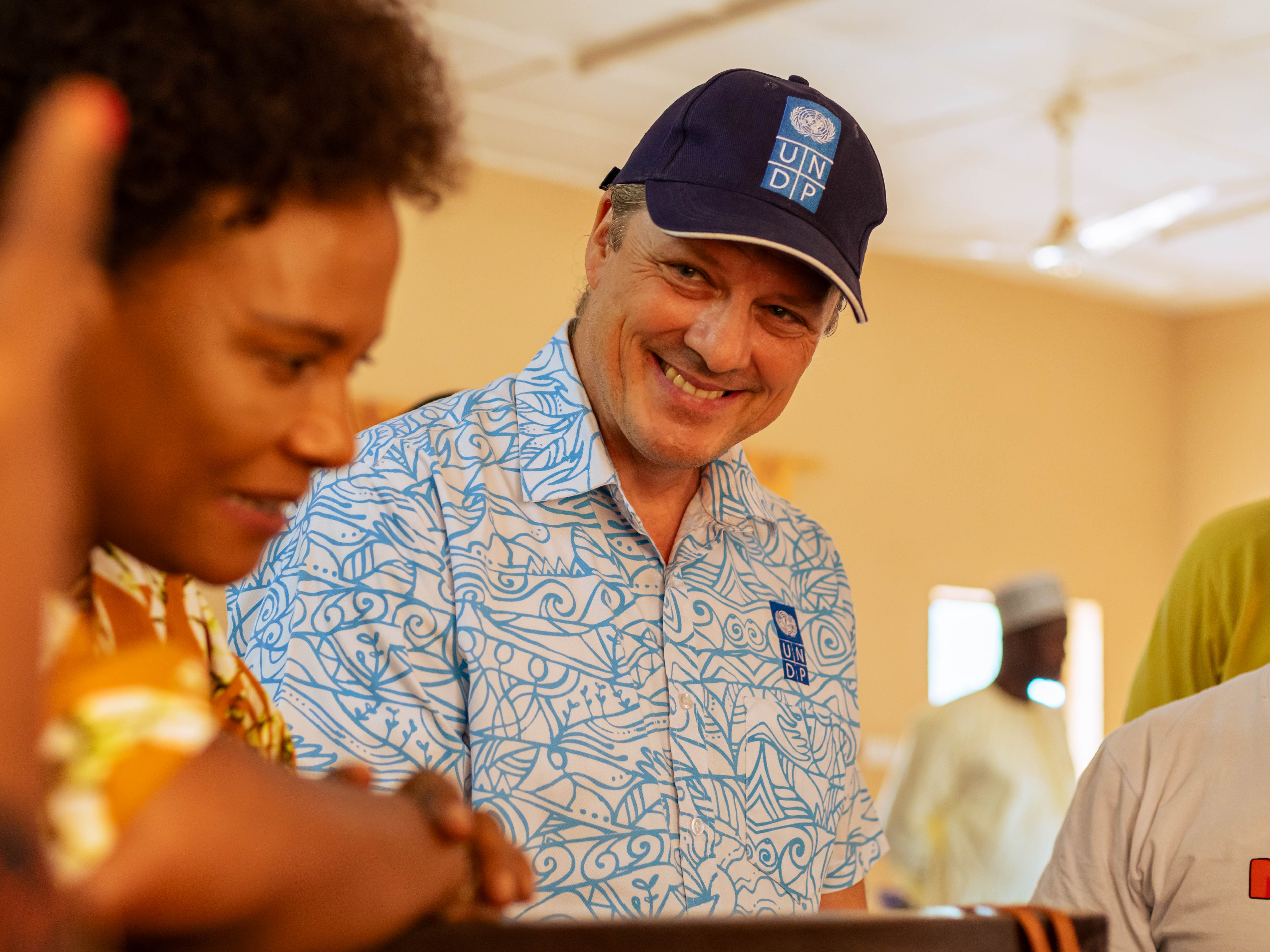 Photo of people around a table; central person in a blue patterned shirt and cap.