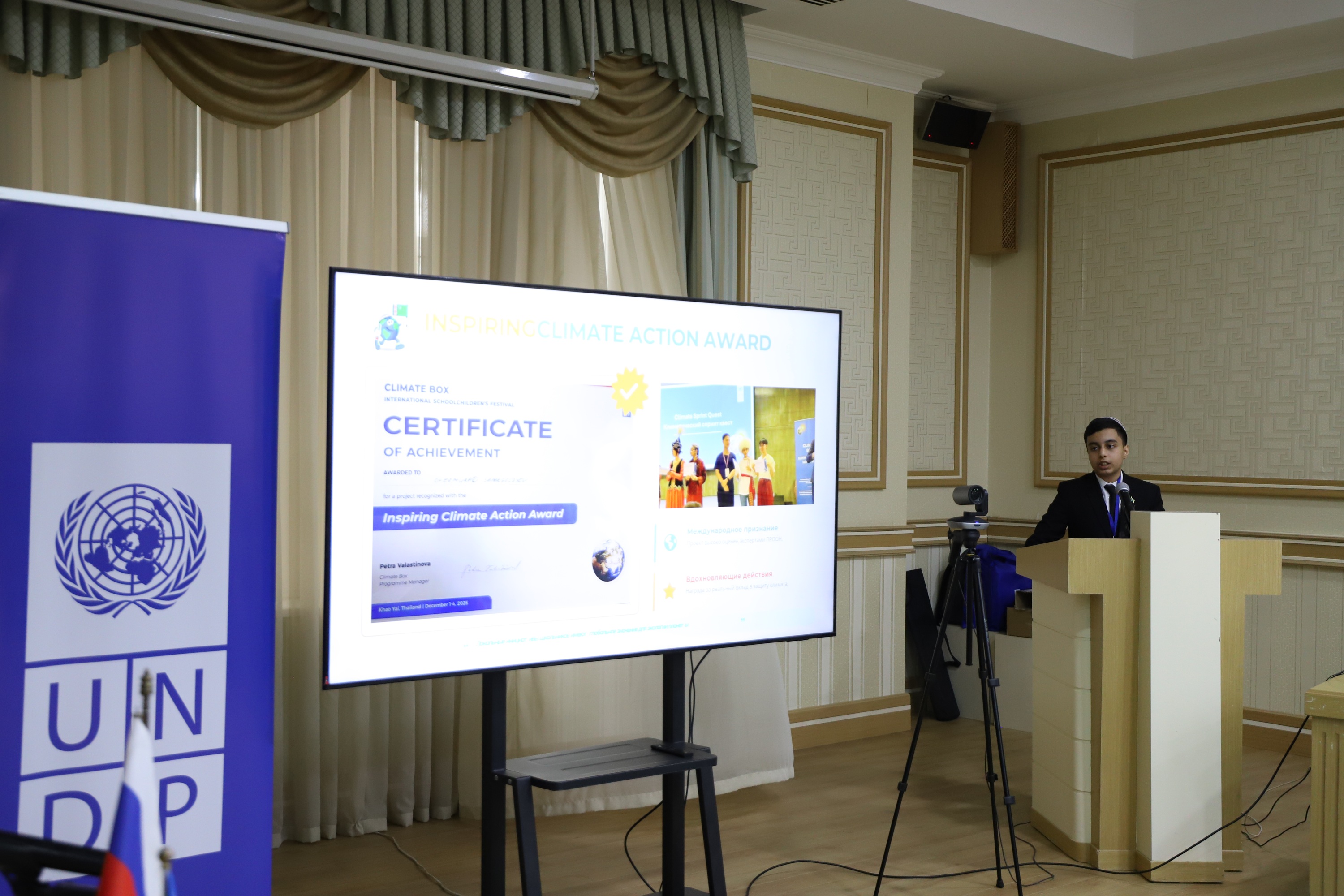 Speaker at a podium presenting slides in a conference room, with a UNDP banner on the left.