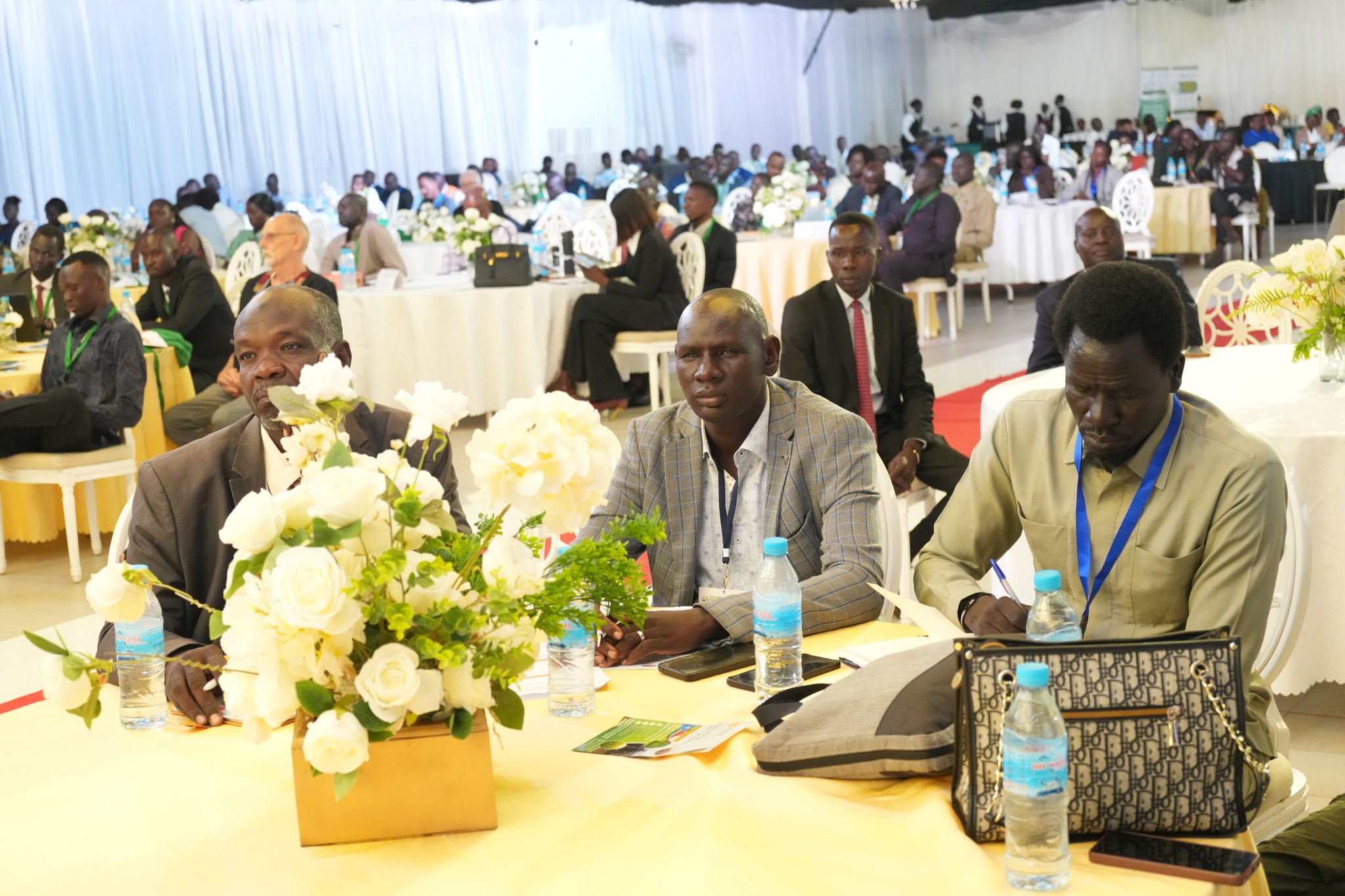 Photograph of a conference hall with many attendees; two men sit at a table in the foreground with a flower centerpiece.