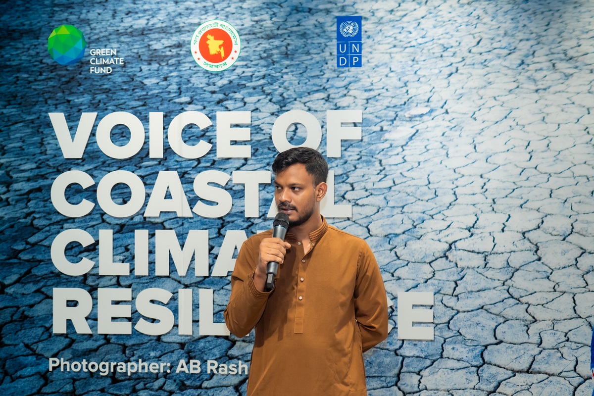 Man in a brown shirt speaks into a microphone on a stage with a blue cracked-earth backdrop.