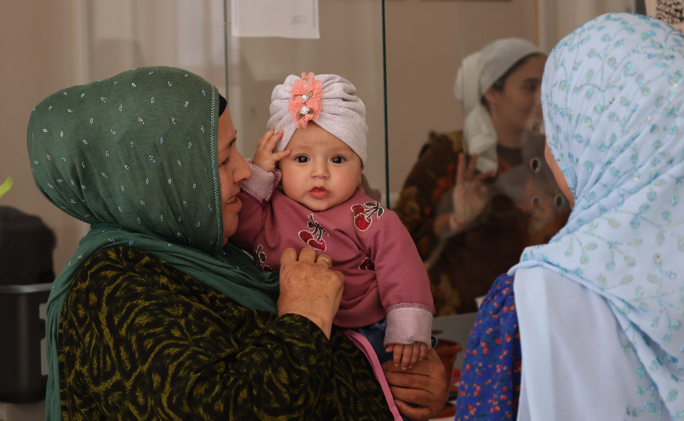 Photograph: woman in green hijab kisses a pink-clad baby; another blue hijab woman nearby.