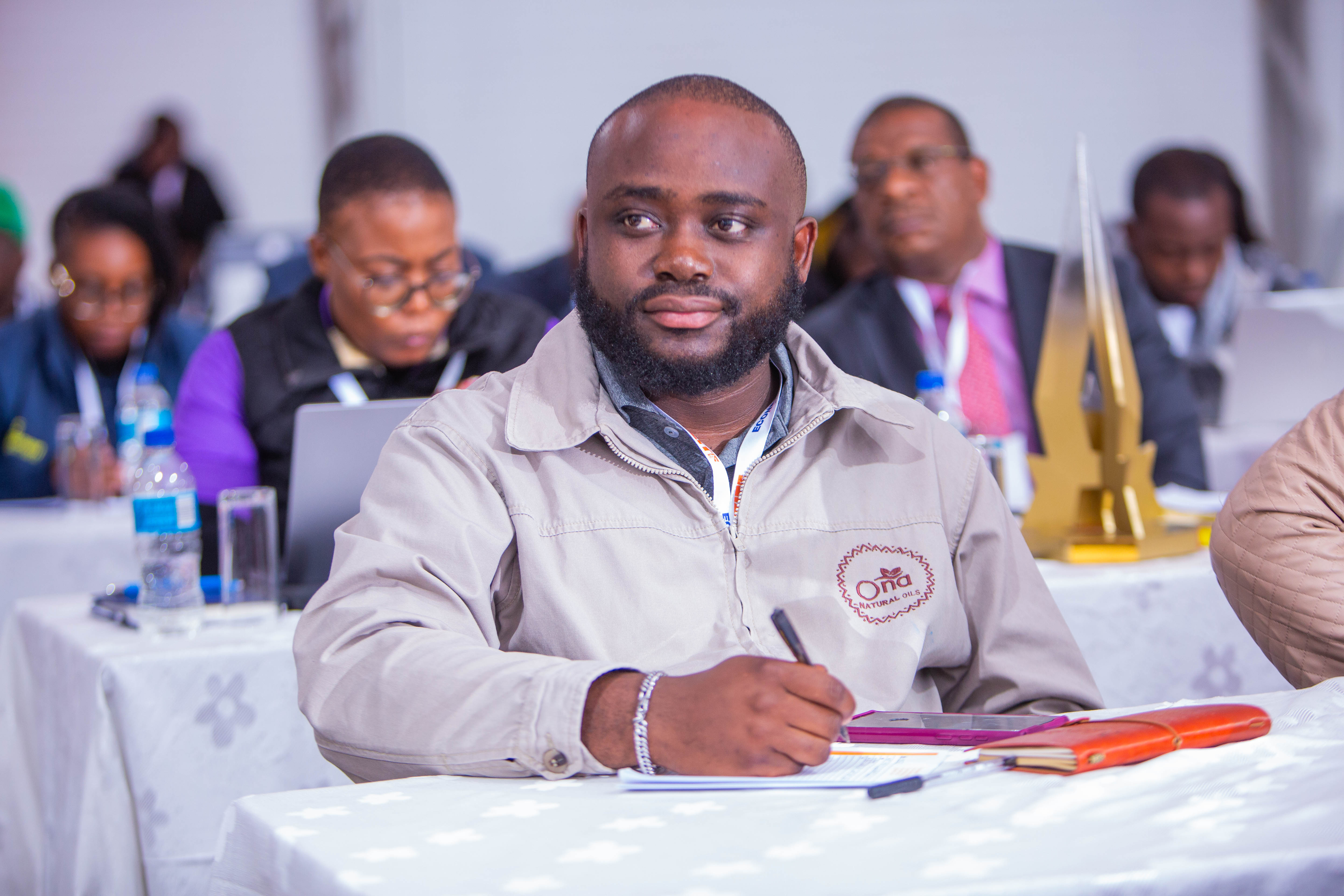 Photograph shows a man in a beige jacket writing at a conference table, with other attendees in the background.