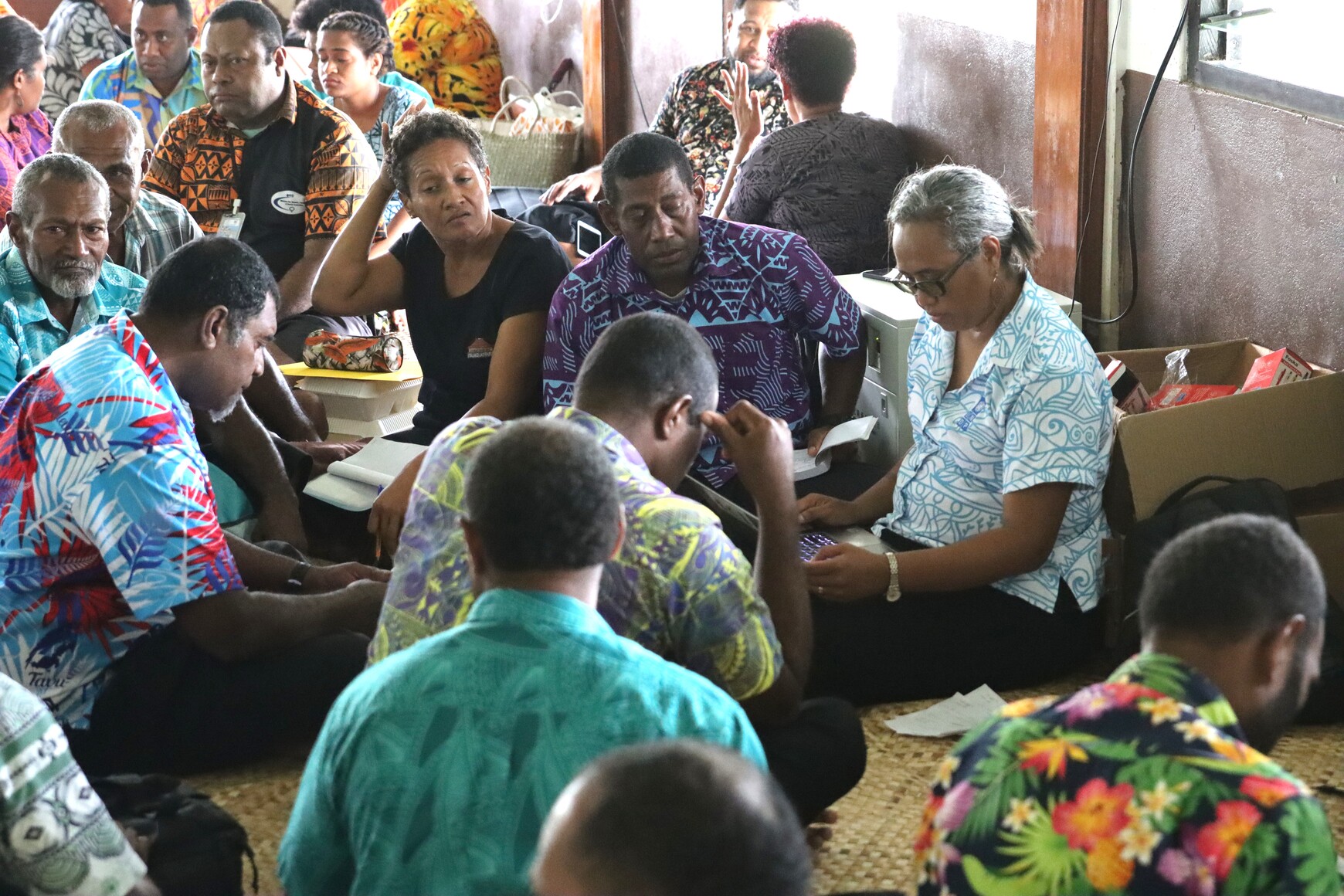Photograph of a crowded room with people in colorful shirts gathered together.
