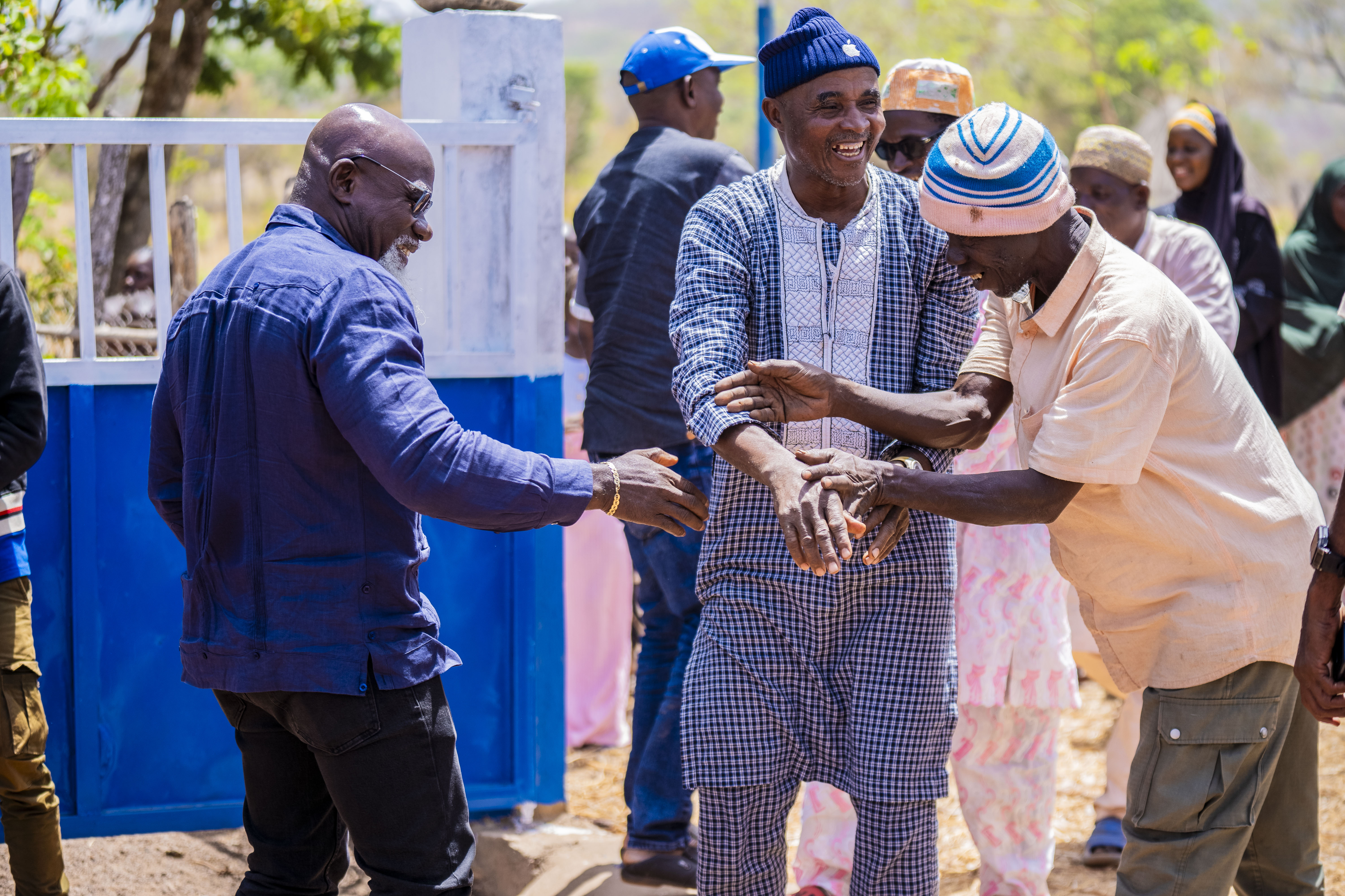 Group of men dancing and smiling outdoors in a sunny courtyard with blue and white walls.