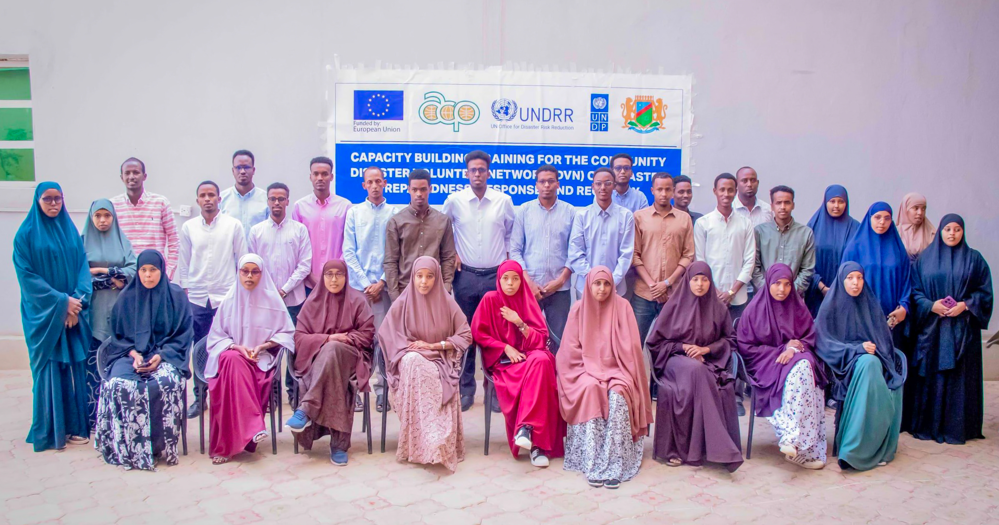 Group in colorful traditional attire posing for a group photo in a hall with a blue banner.