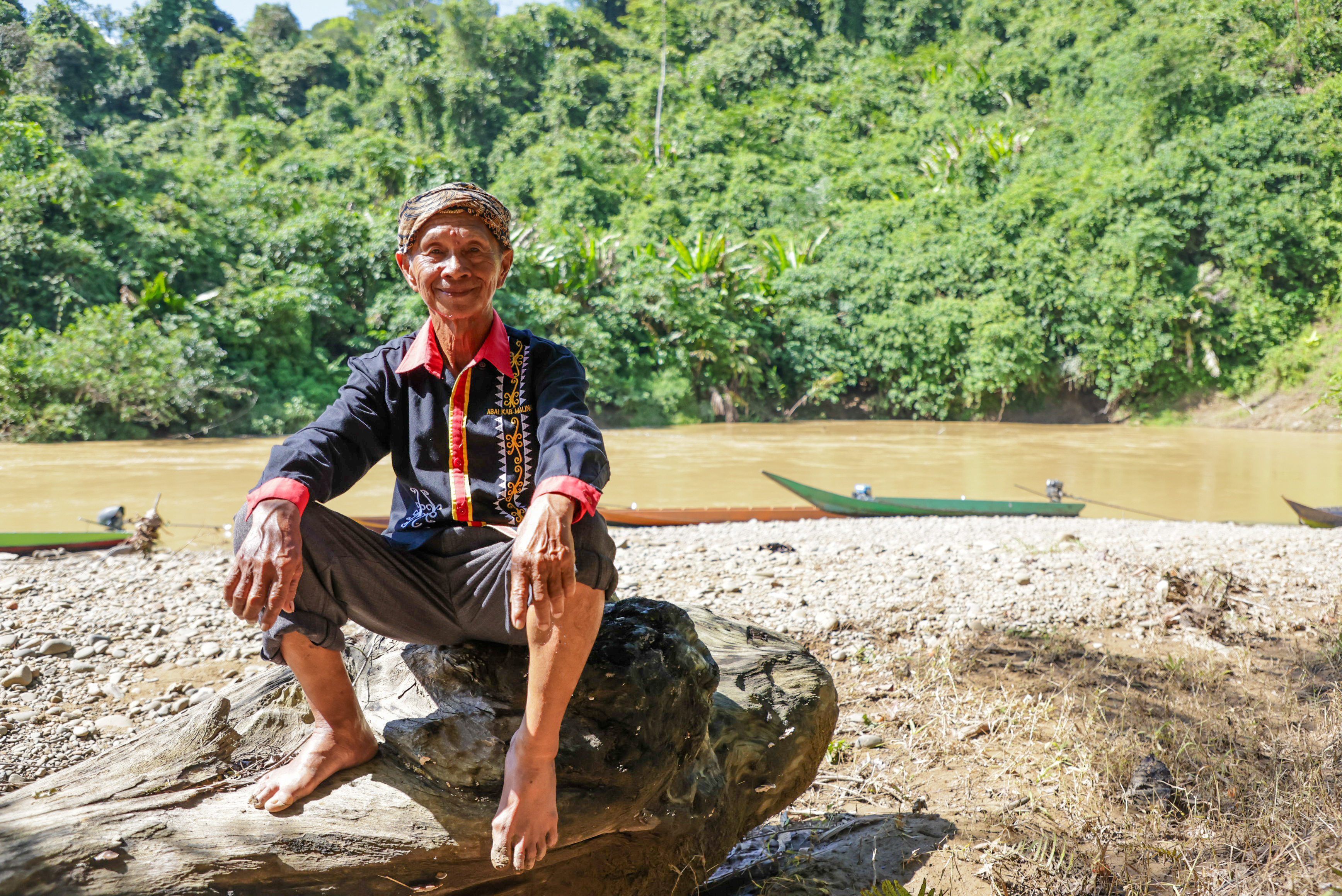 Photo of a man sitting on a rock by a river, with green forested hills in the background.
