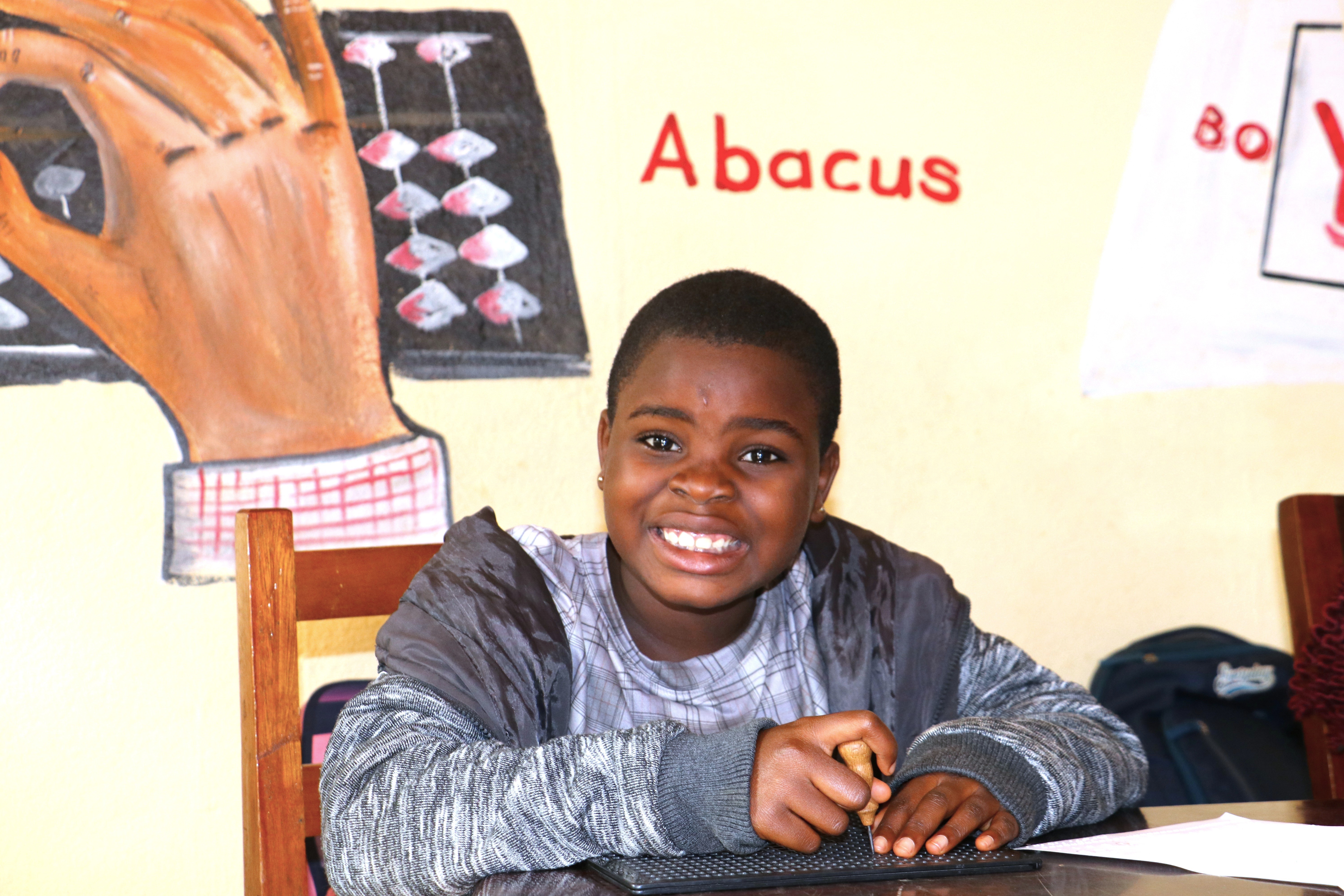 Photograph of a student with a blurred face at a desk in a classroom, abacus posters on wall.