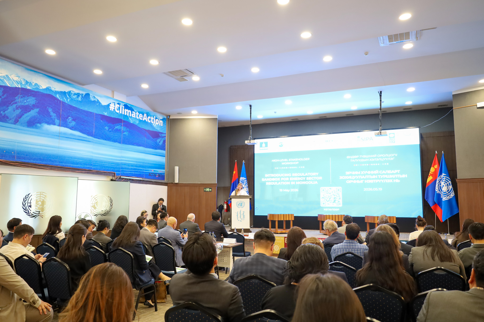 Conference room full of attendees facing a speaker at a podium with a large screen and flags.