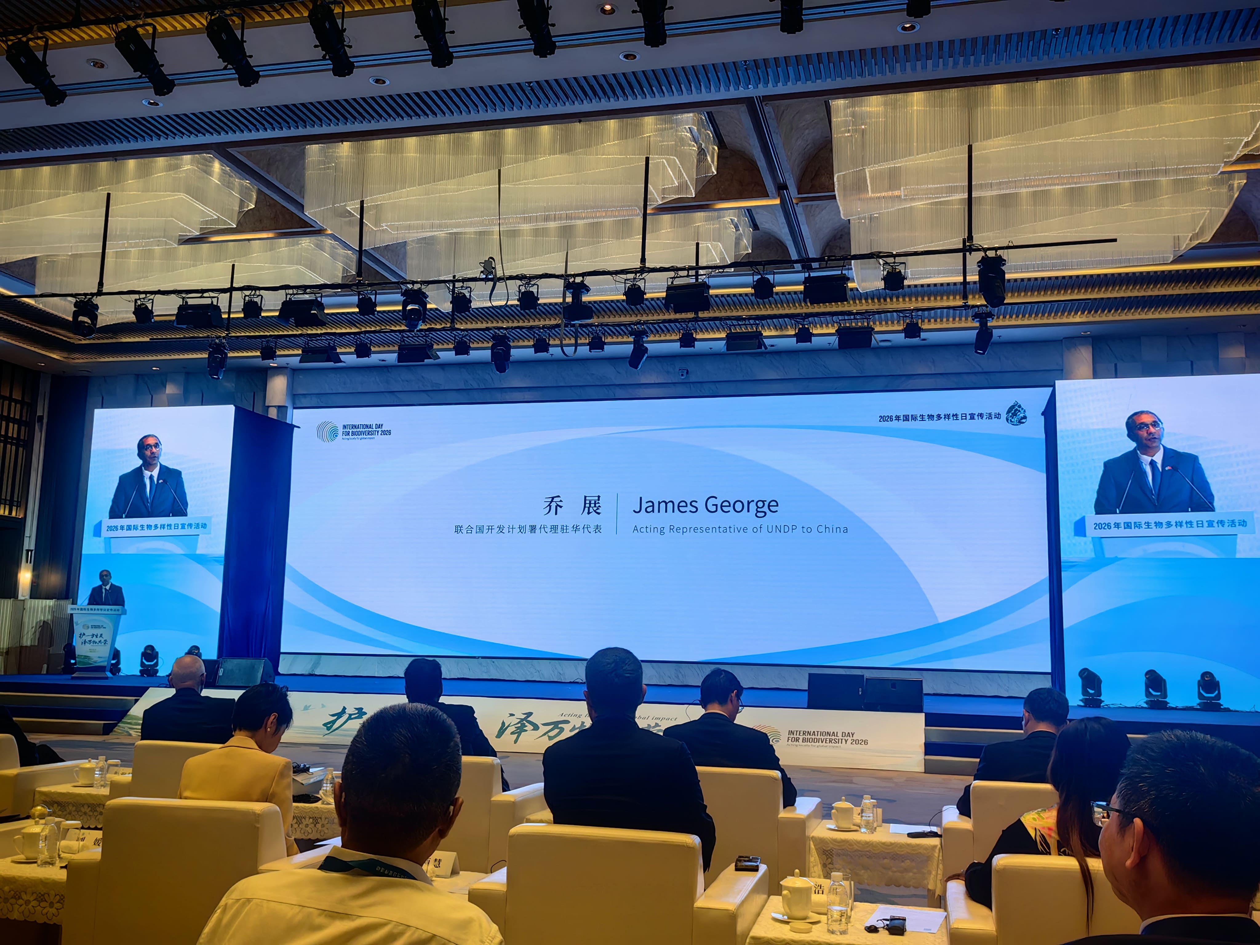 Conference hall with a blue-lit stage, three large screens, and attendees seated at tables.