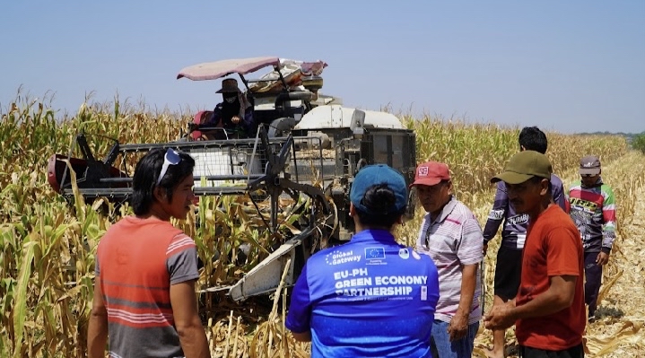 People stand in a cornfield around a tall trailer-mounted harvester under a blue sky.