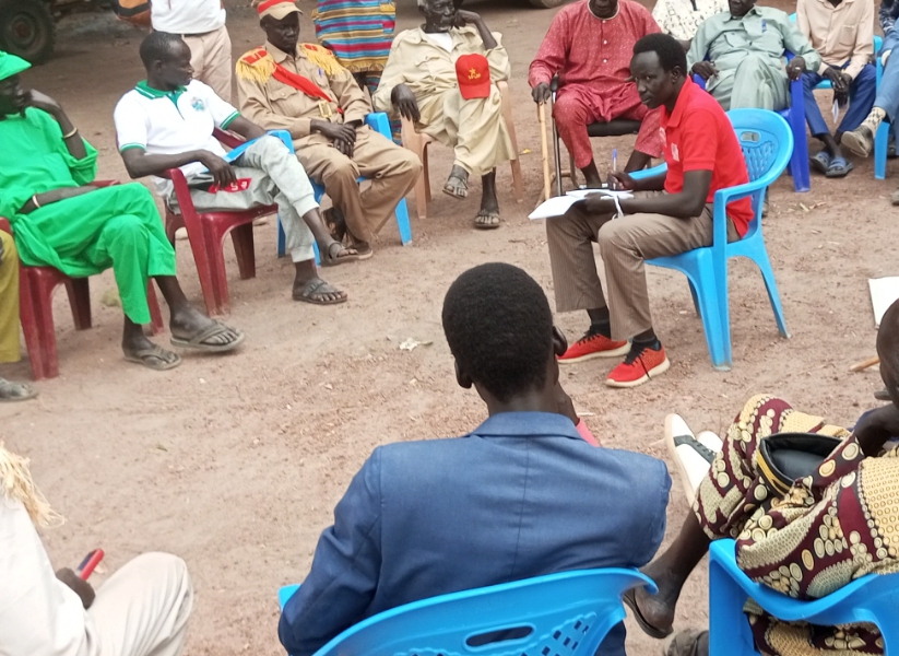Group of people in colorful clothes sit in a circle on plastic chairs outdoors on dirt ground.