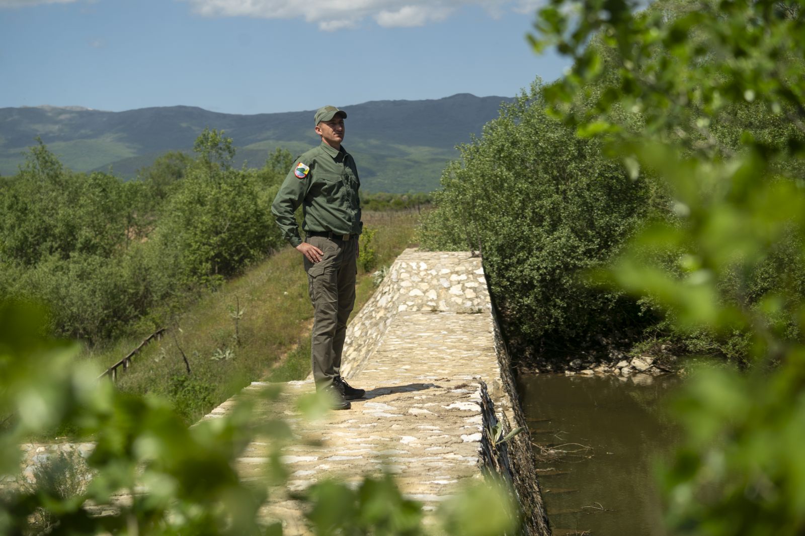 Man in dark jacket stands on a stone footbridge over a narrow stream, with hills in the distance.