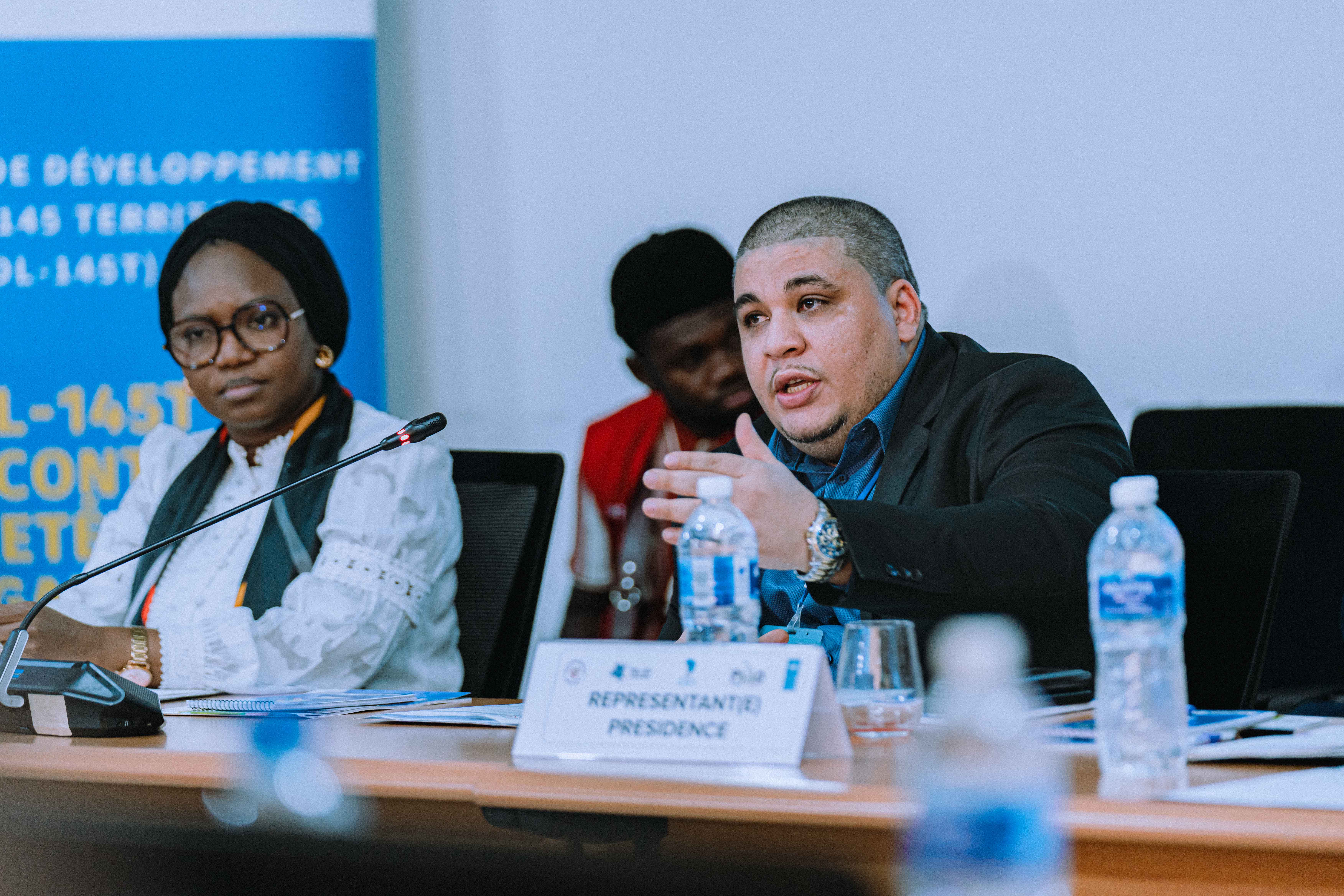 Attendees at a conference table; water bottles, a laptop, blue banner in background.