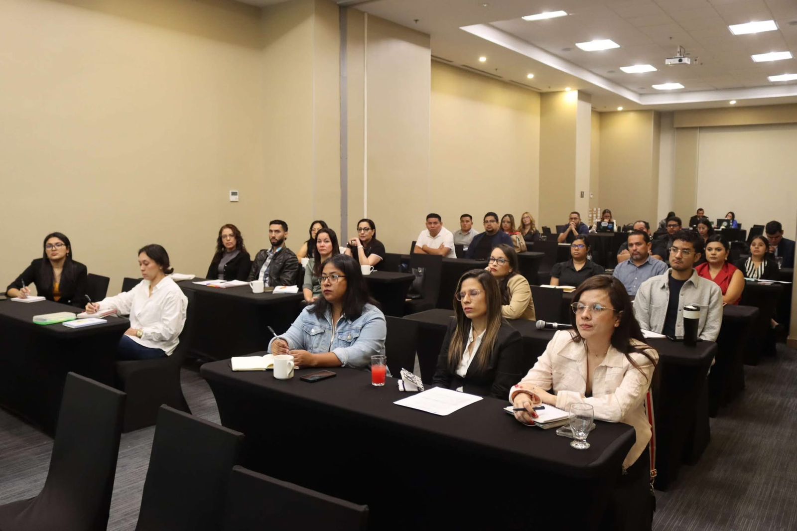 Audience seated in a conference room, rows of desks with name placards as attendees listen.