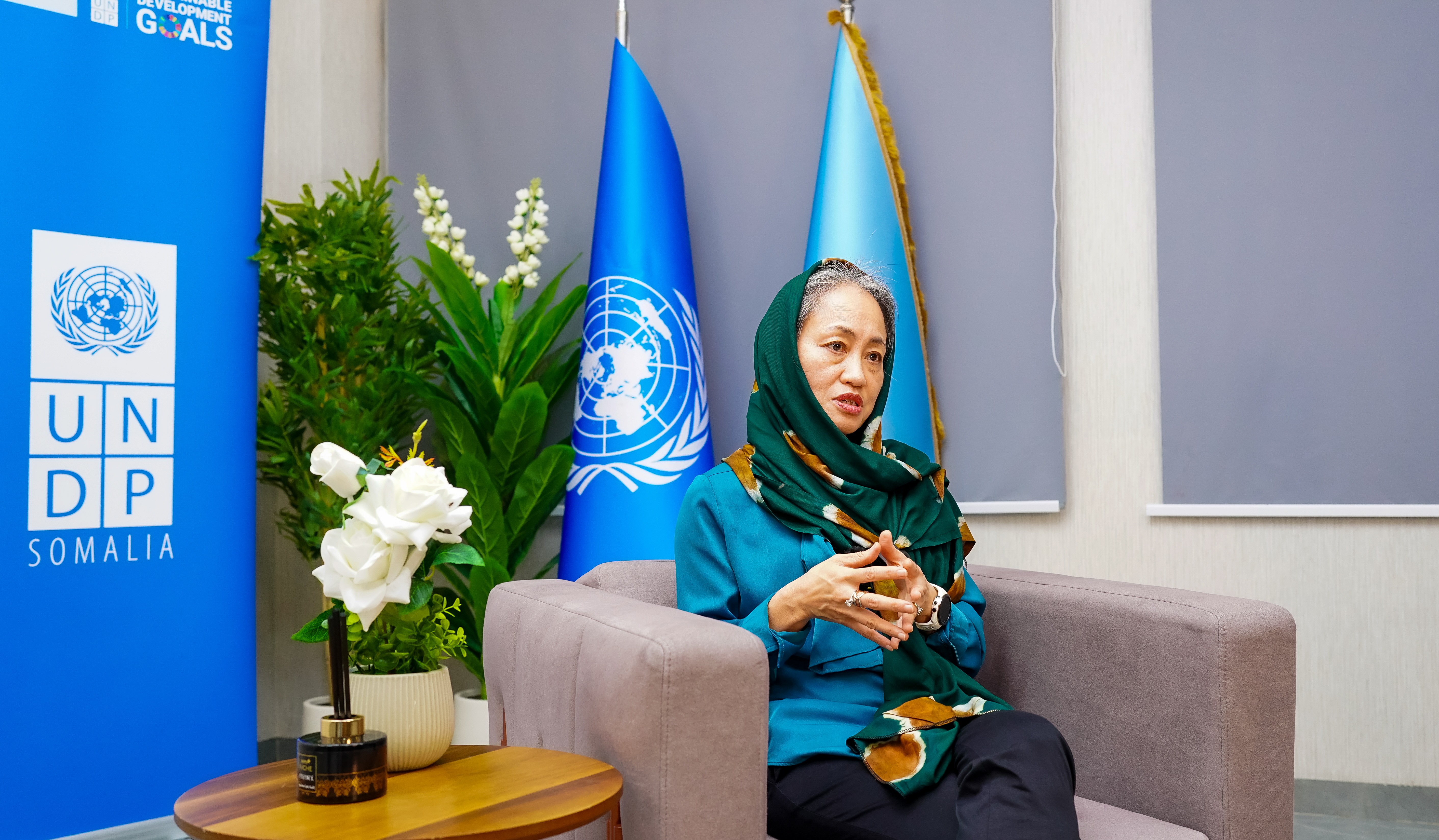 Woman in hijab seated in a UN room with blue flags and UNDP banner.