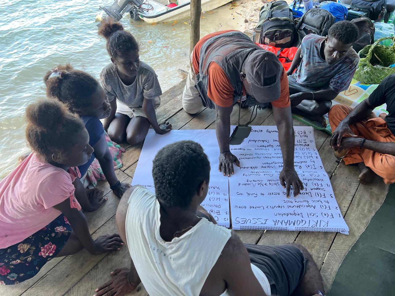 Group on a wooden dock by the water, gathered around a large handwritten sheet.