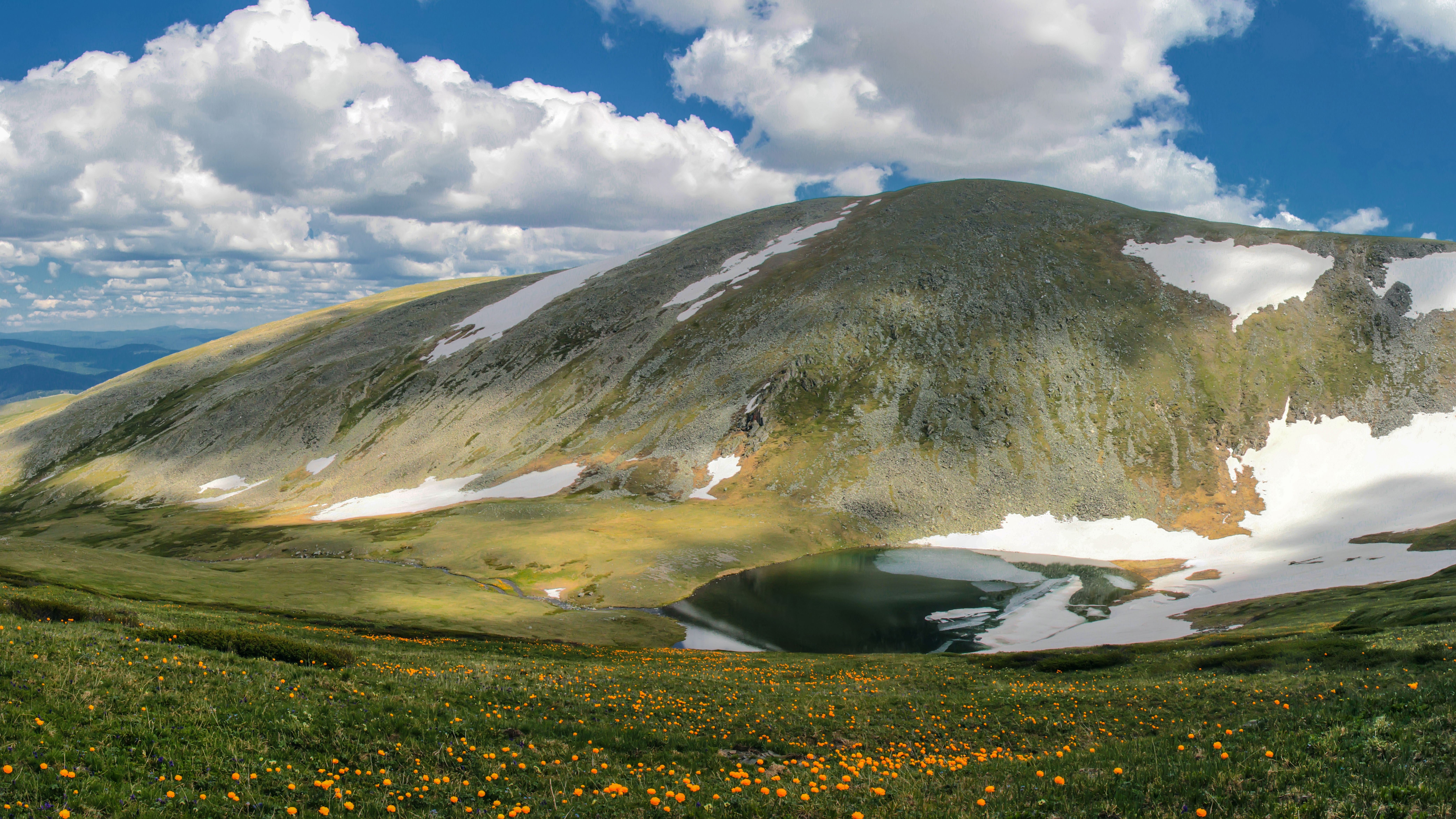 Alpine meadow with yellow flowers, green slopes, and a caldera lake under a blue, cloudy sky.