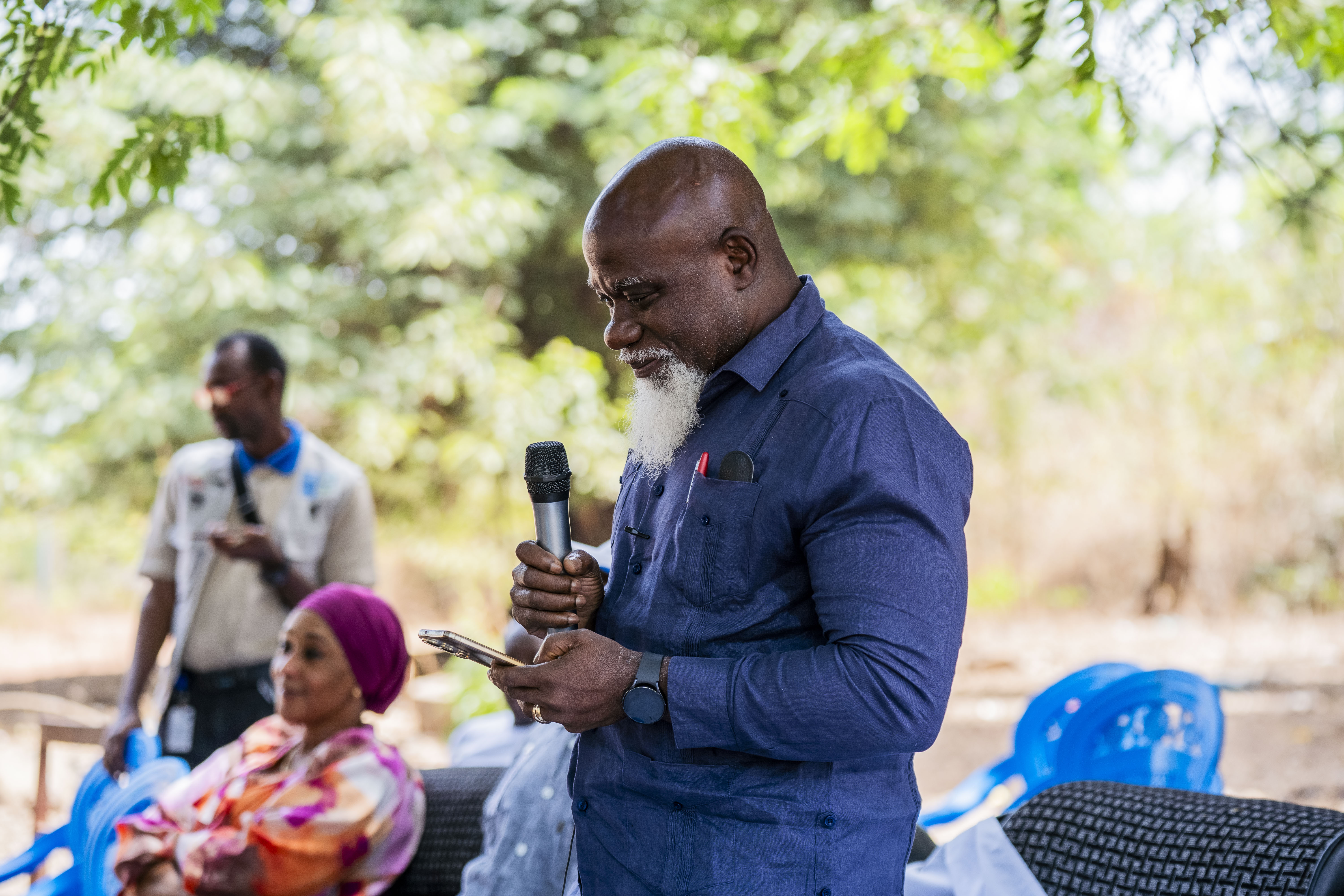Man in blue shirt speaks into a microphone at an outdoor event; seated audience.