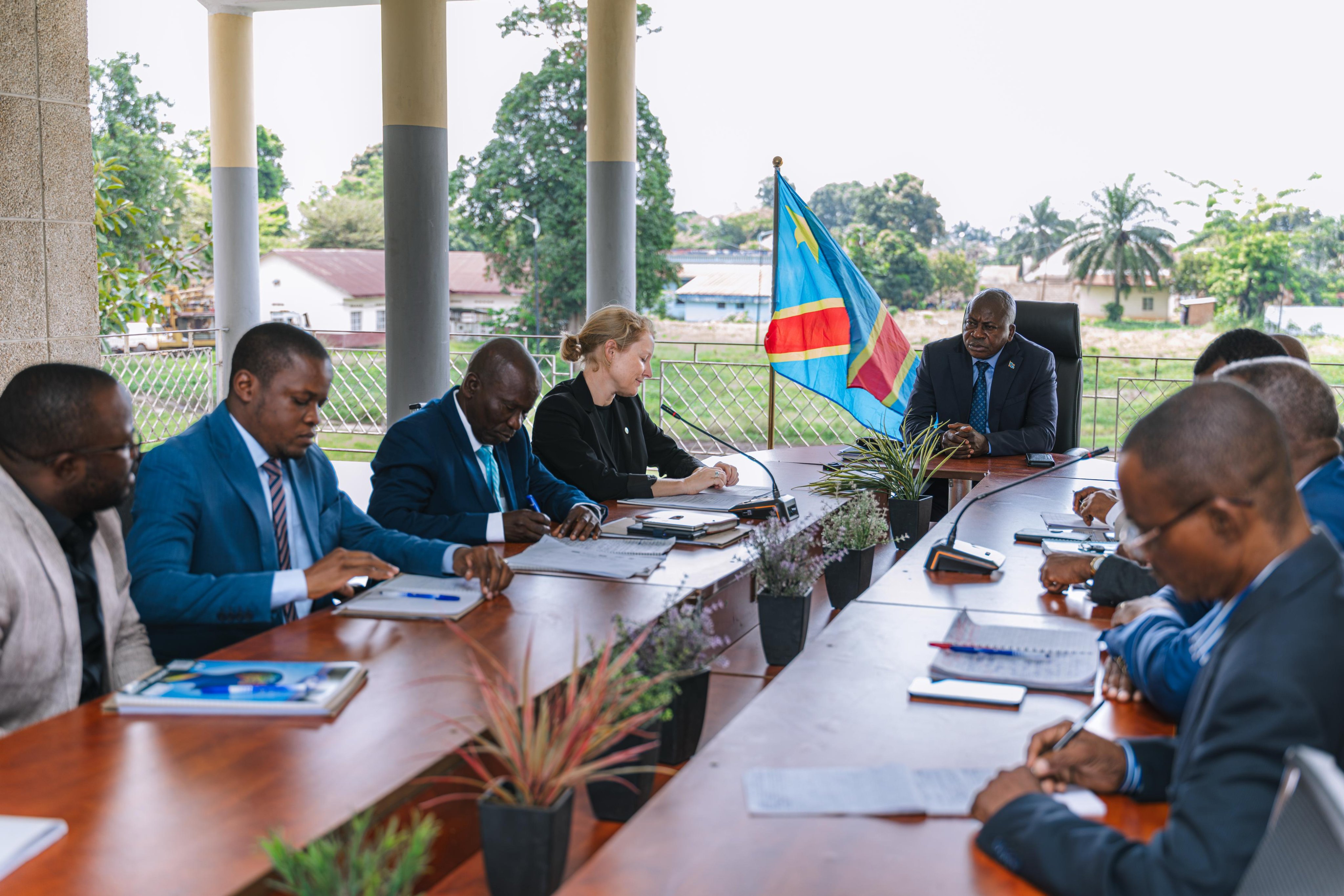 Group of professionals in business attire around a long conference table on a veranda; documents on the table.