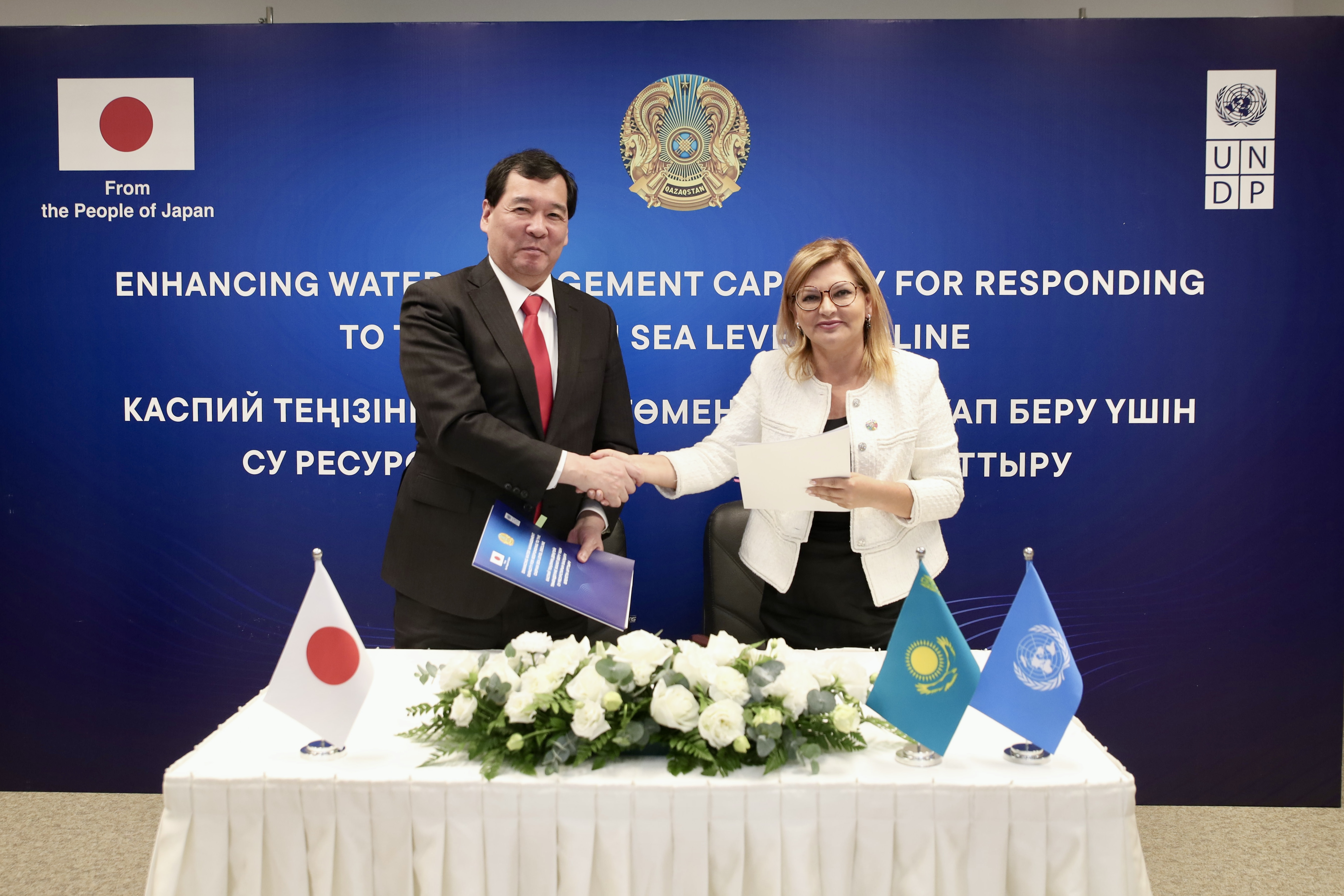 Two men in suits shake hands across a table during a signing ceremony; Japan and Kazakh flags.