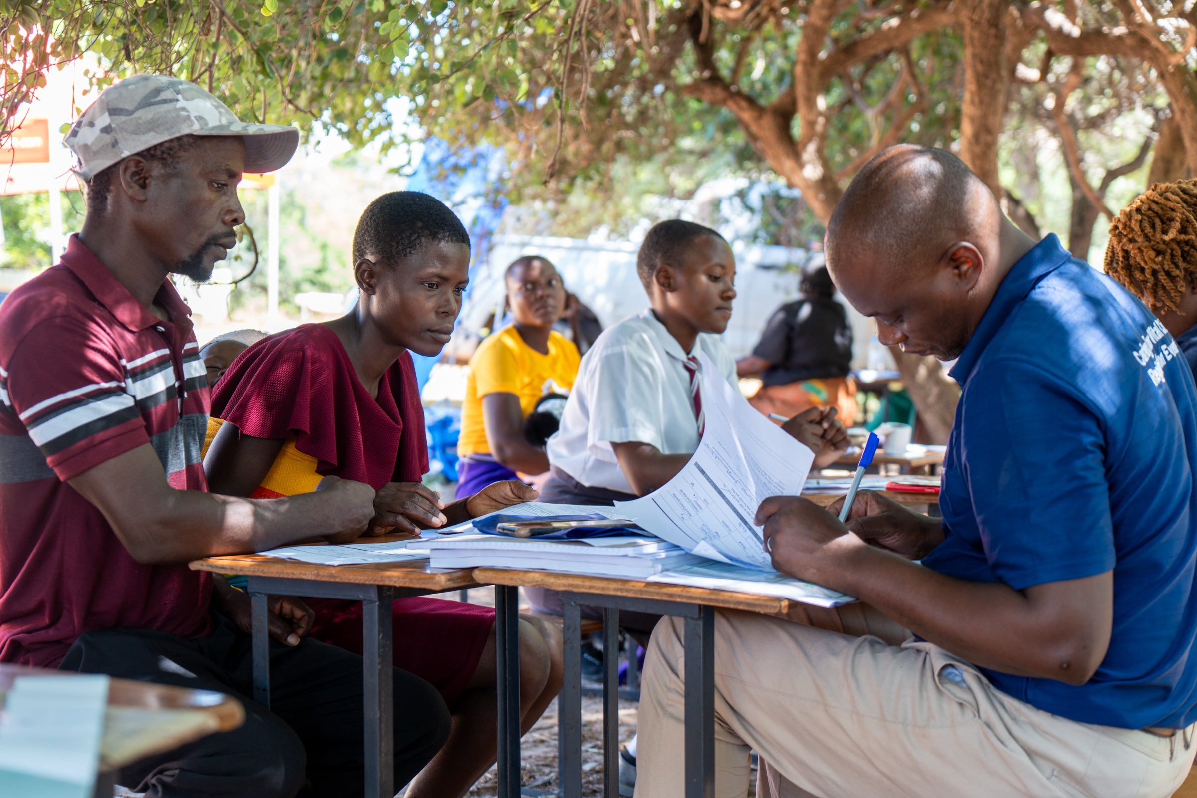 Outdoor classroom with diverse people seated at a long table, writing on papers under trees.