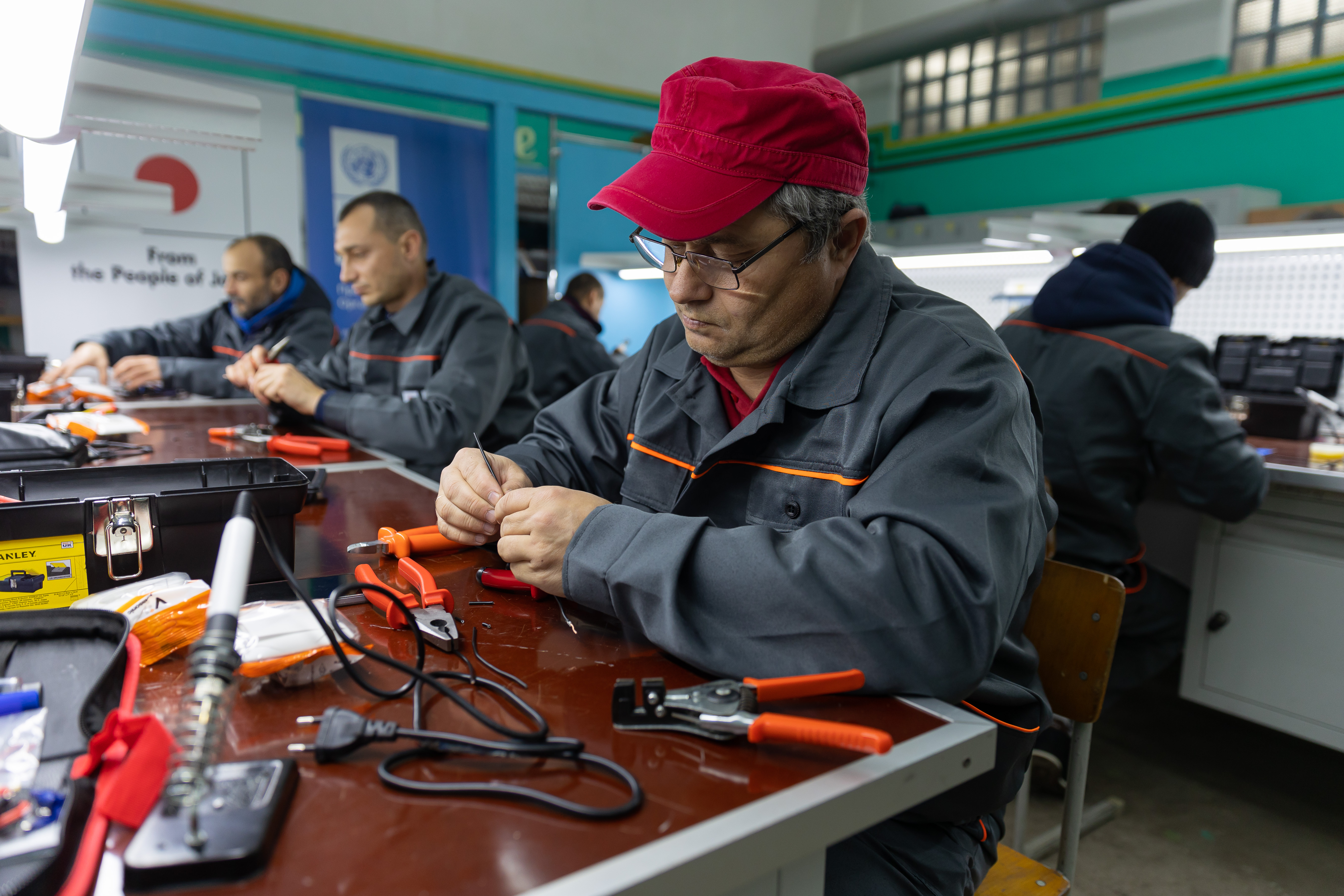 Worker with a red turban soldering at a bench in a busy electronics workshop; others work.