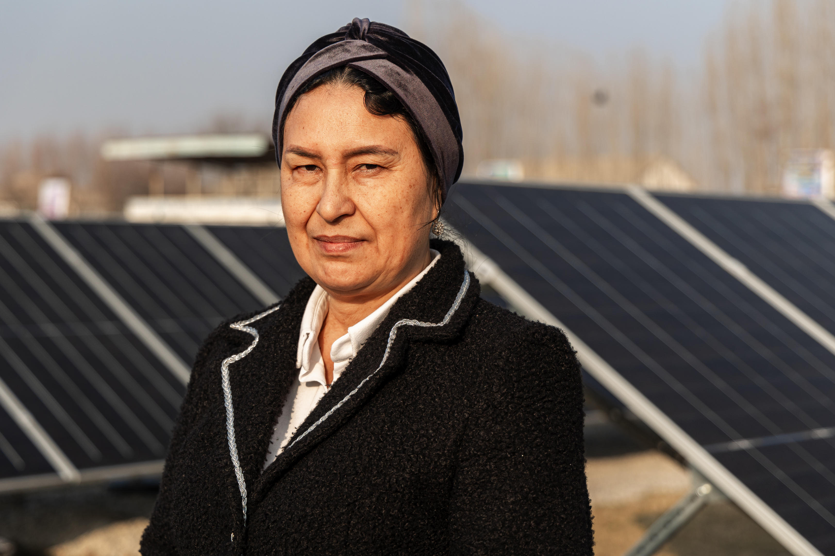 Person in a dark coat with a fur collar stands near solar panels; face blurred.