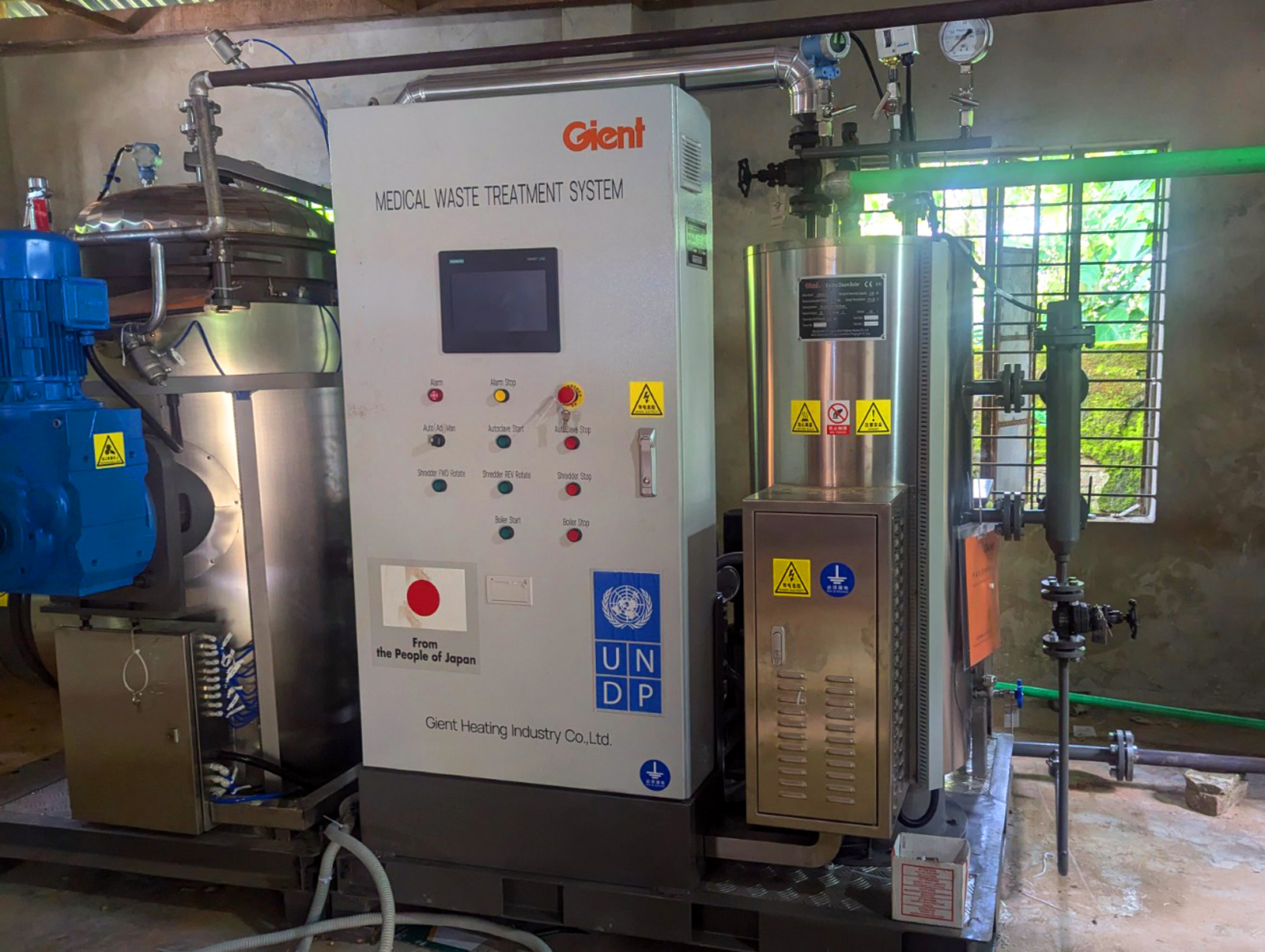 Industrial control panel with touchscreen, red stop button, and blue piping in a mechanical room.