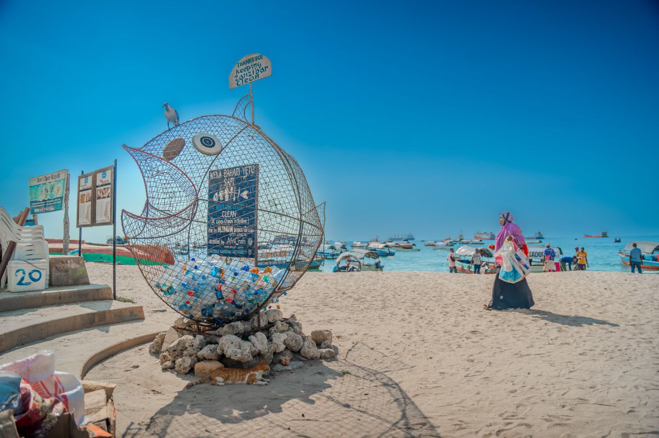 Blue-and-white mosaic round sea-creature sculpture on a sandy beach with a chalkboard sign.