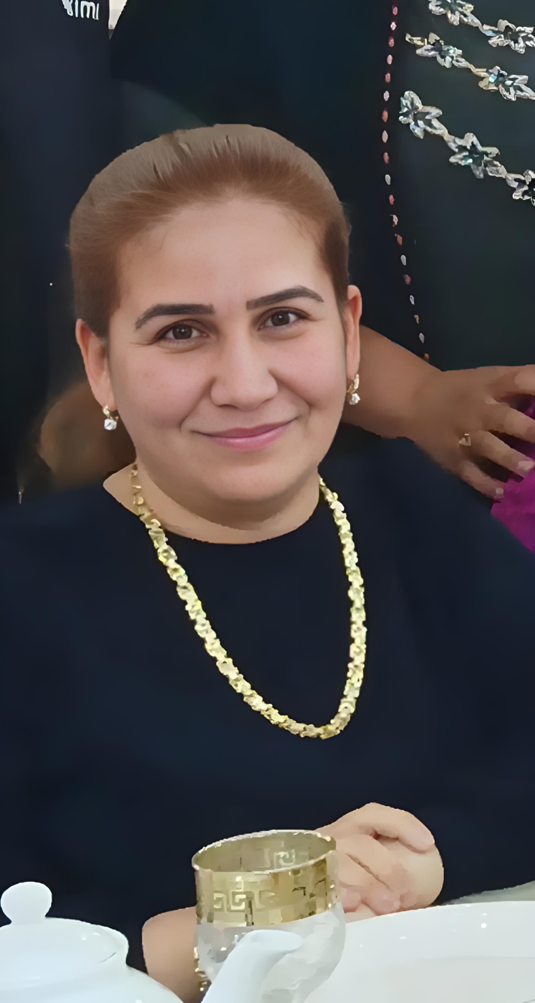 Person with blurred face wearing a gold chain and dark top, seated at a table with a teacup.