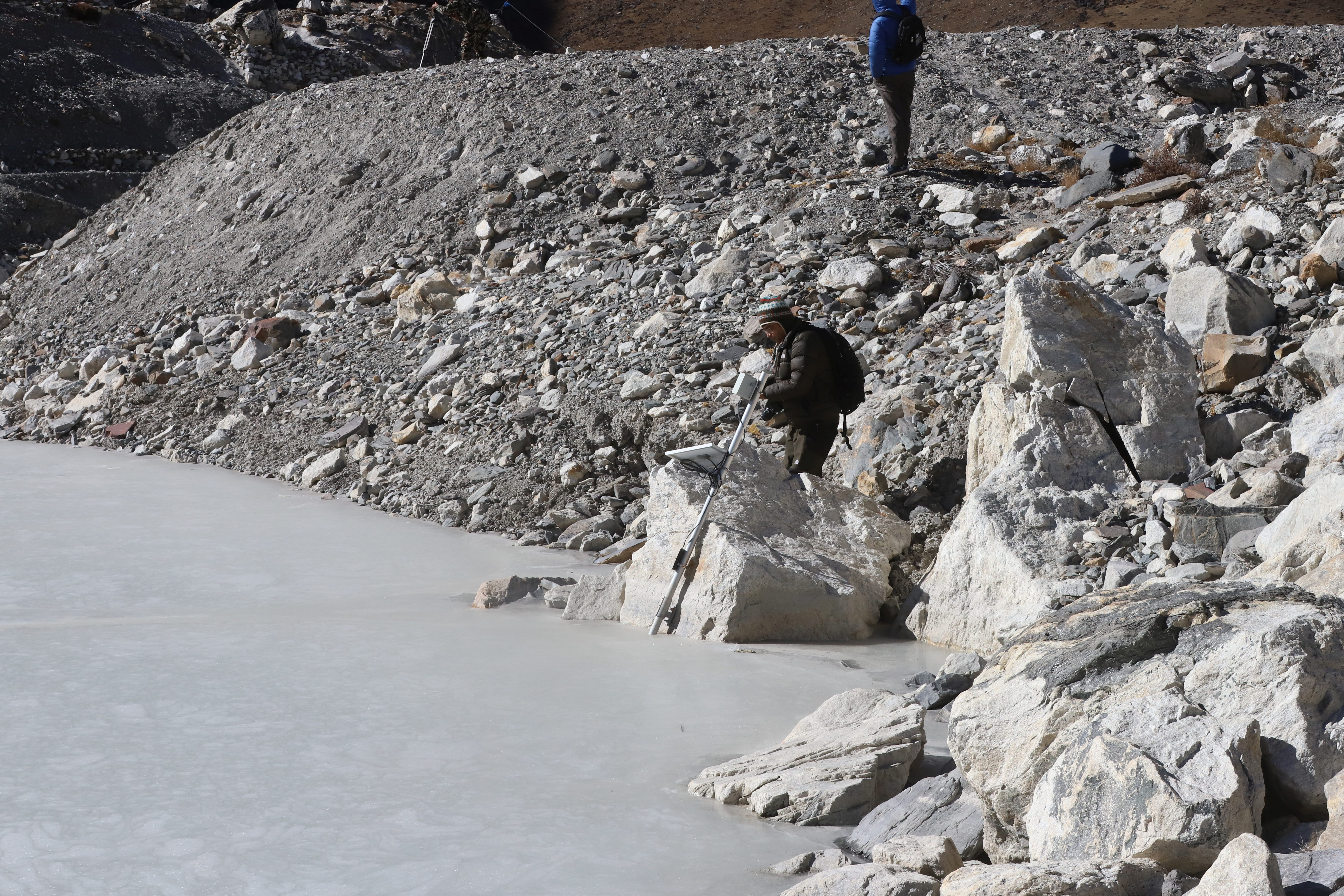 Person standing on a rocky slope above a pale blue icy surface near a cave opening.