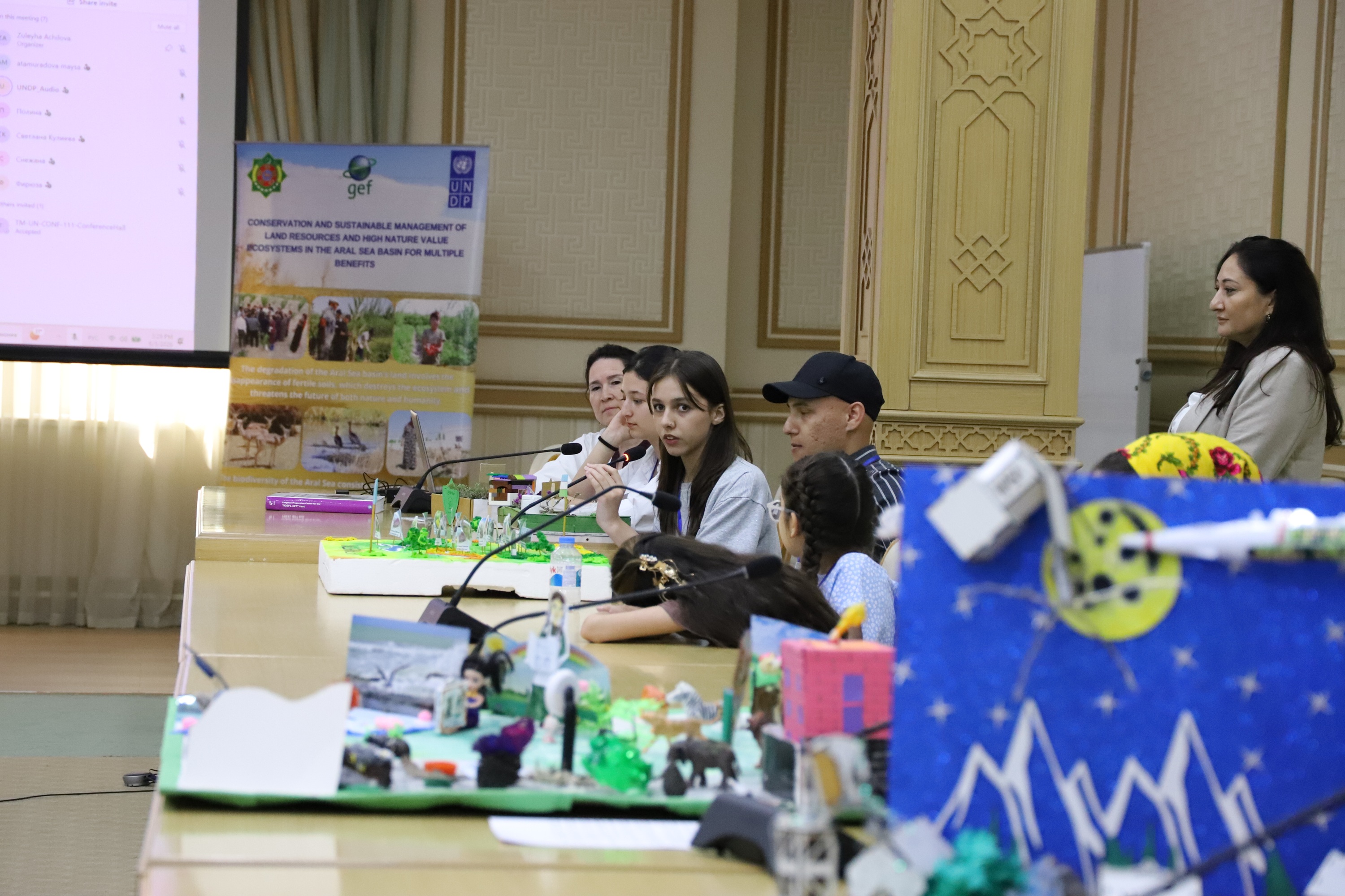 Speakers' panel at a long table; presenter stands to the right, slides on screen.