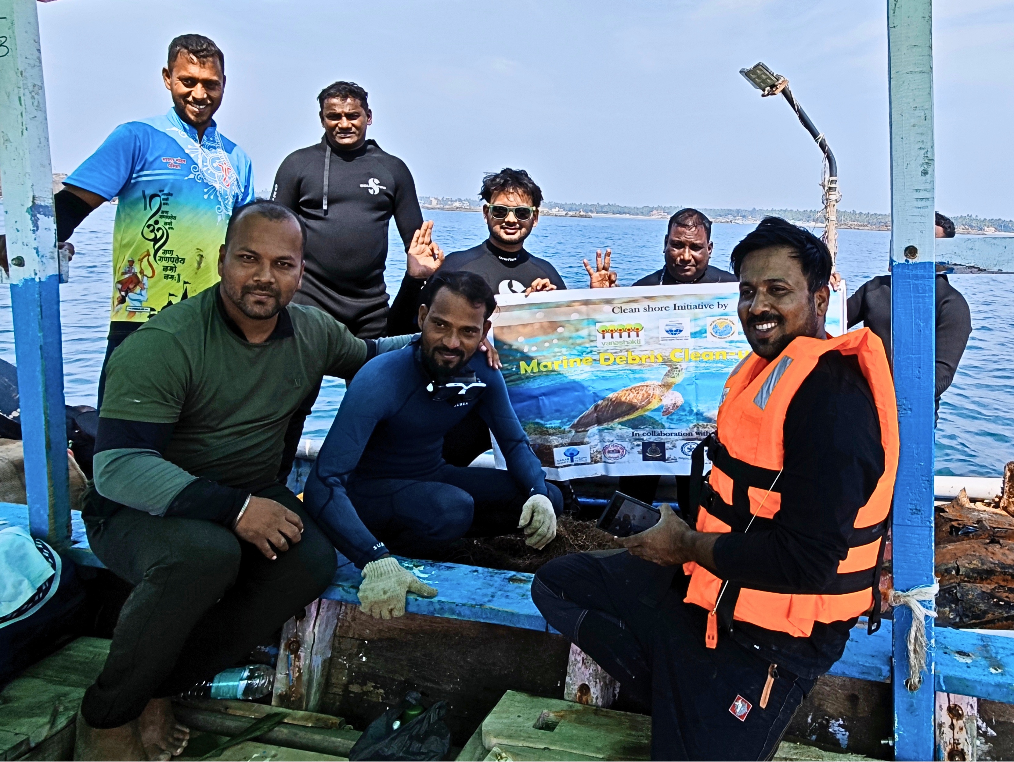Group of divers in wetsuits and life jackets on a blue boat, posing with a map sign by rocky shore.