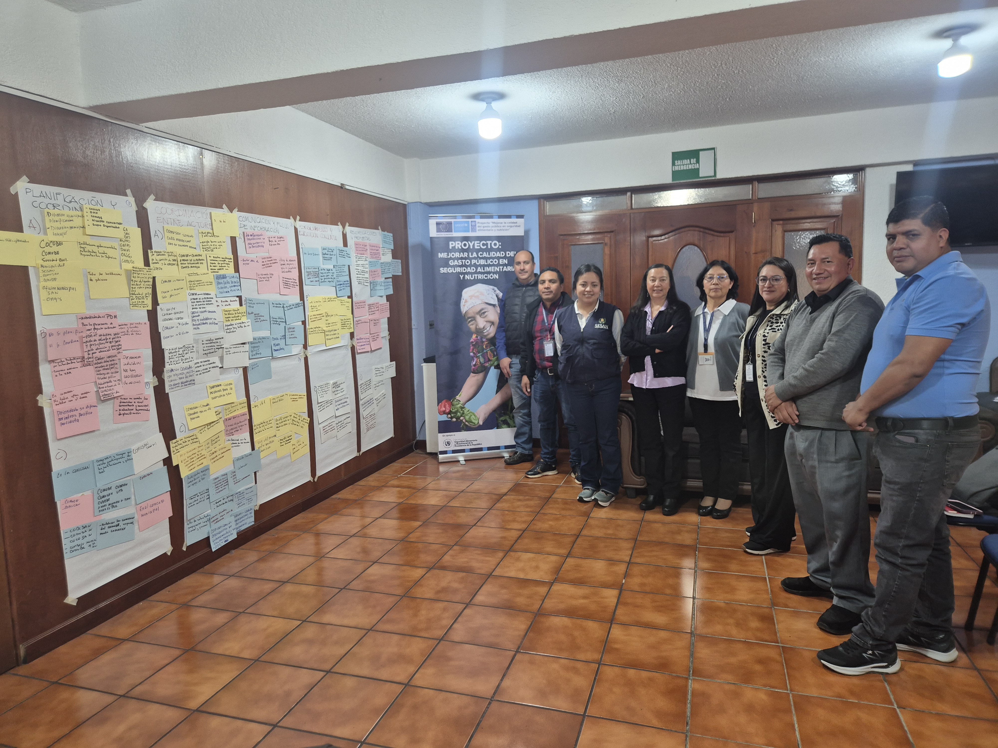 Group of people posing in a hallway with a wall covered in colorful sticky notes and posters.