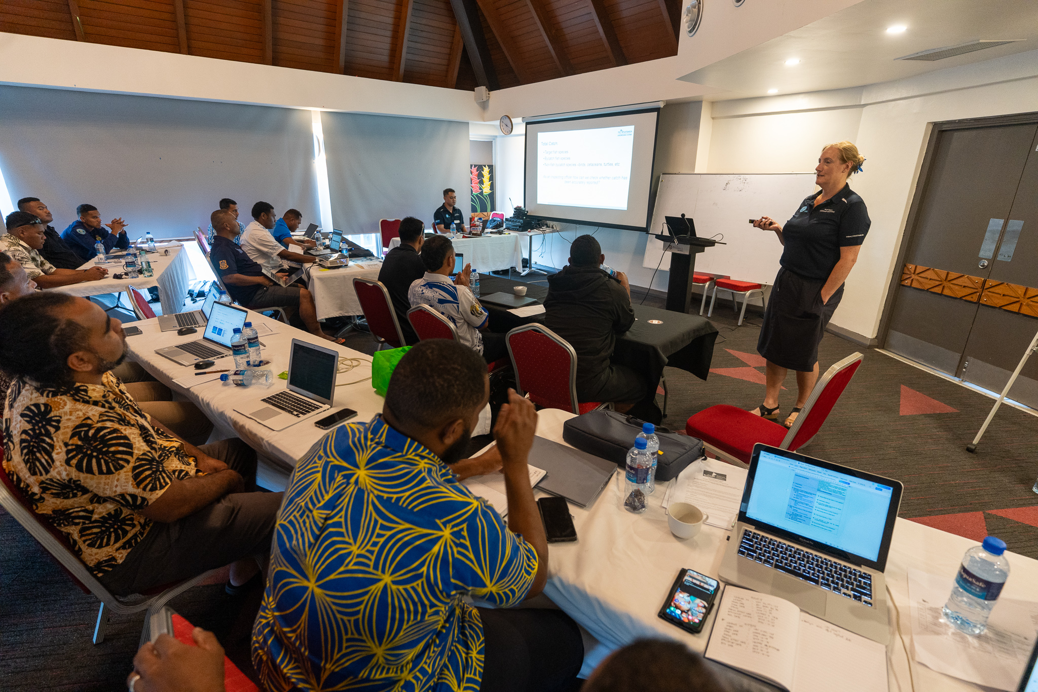 Photo of a workshop: attendees around tables while a presenter speaks at the front with a screen.