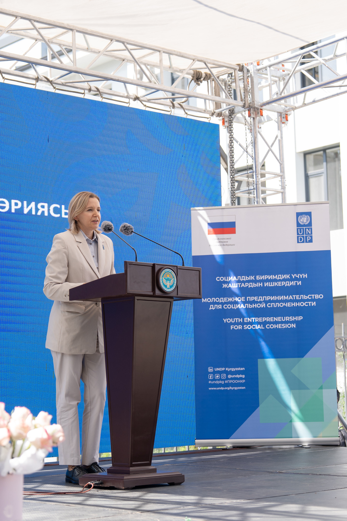 A woman in a light beige suit speaks at a podium on an outdoor stage with blue banners.
