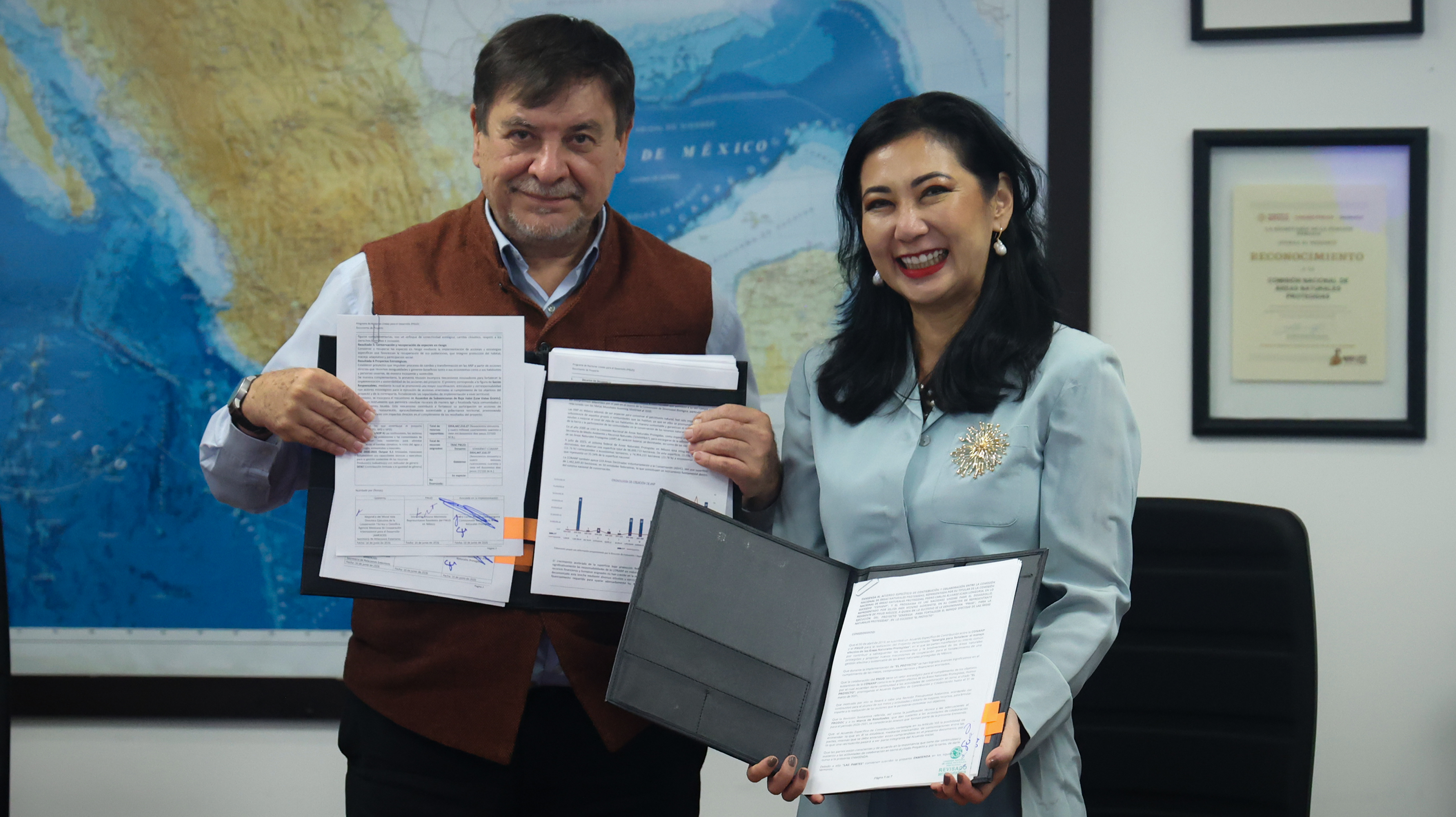 Two people indoors, holding certificates and folders; blue world map and frames on the wall.