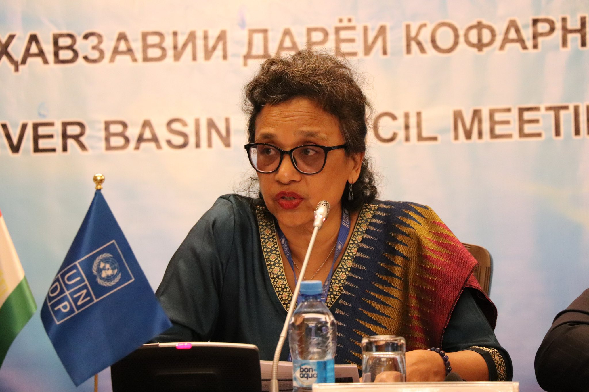 Person with colorful scarf and badge seated at a conference table; blurred face.
