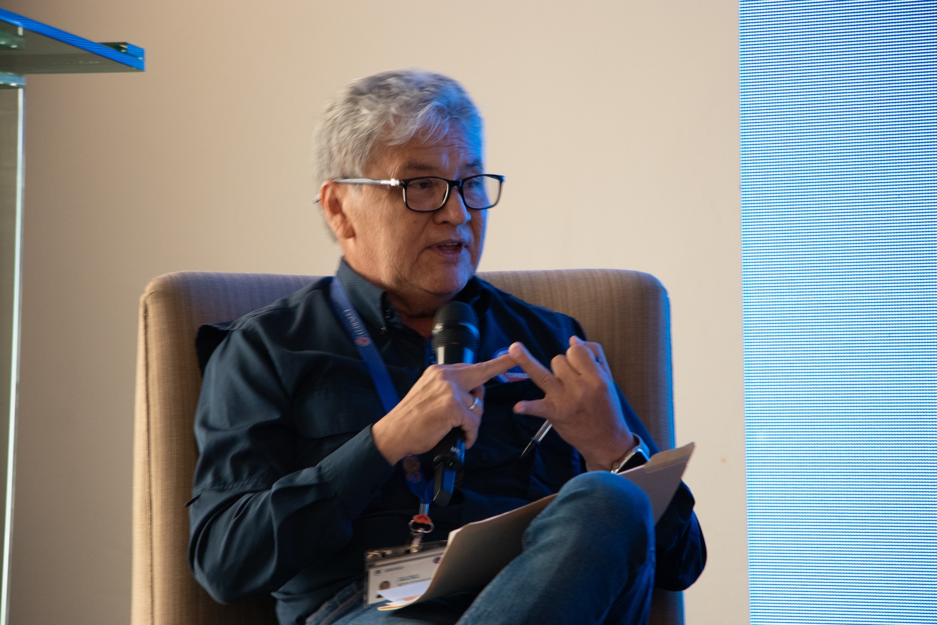 Person with gray hair and glasses sits in a chair, gesturing with hands, wearing a badge.