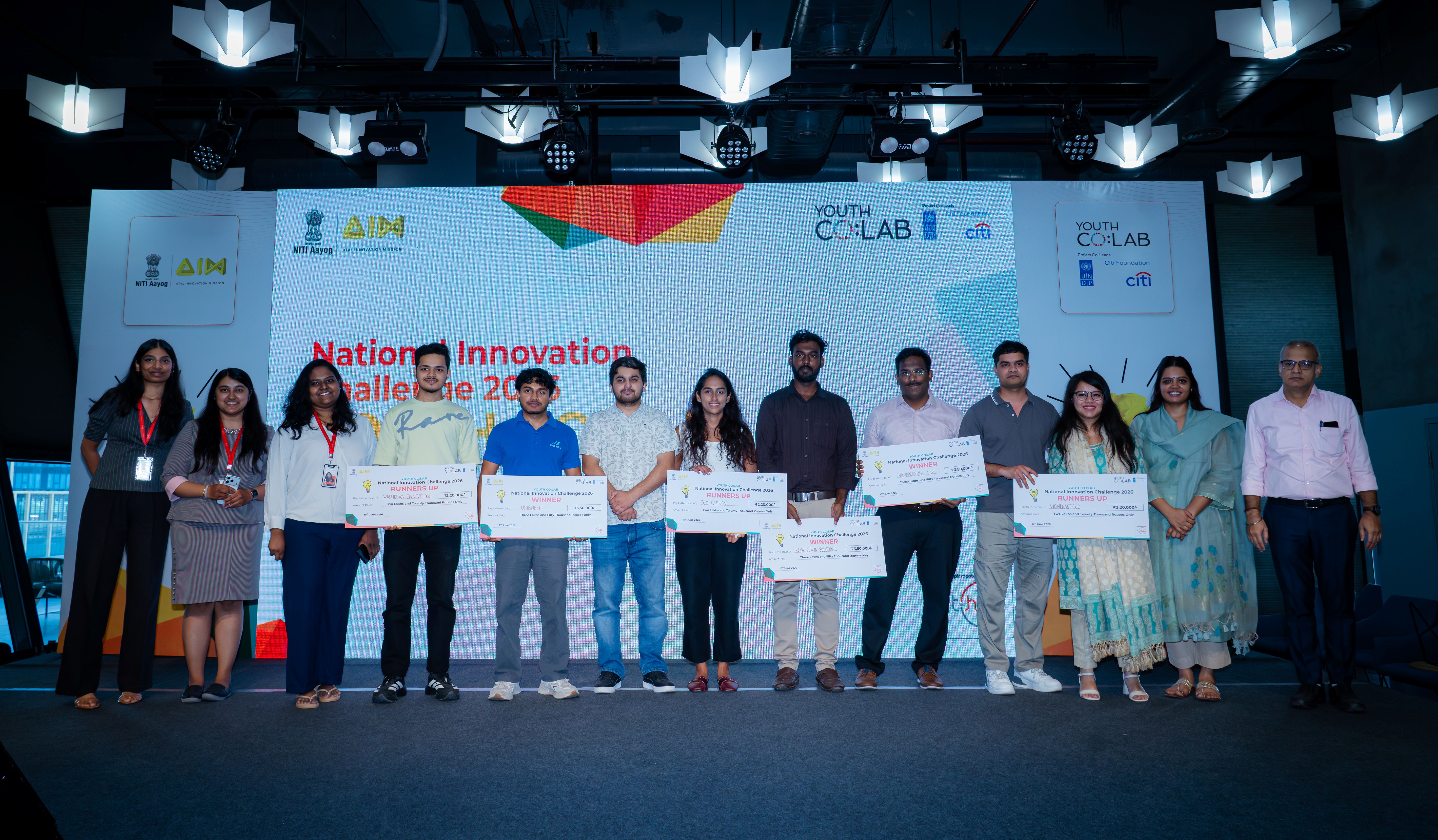 Group of people on a stage at a ceremony, holding certificates, with a blue backdrop featuring logos.