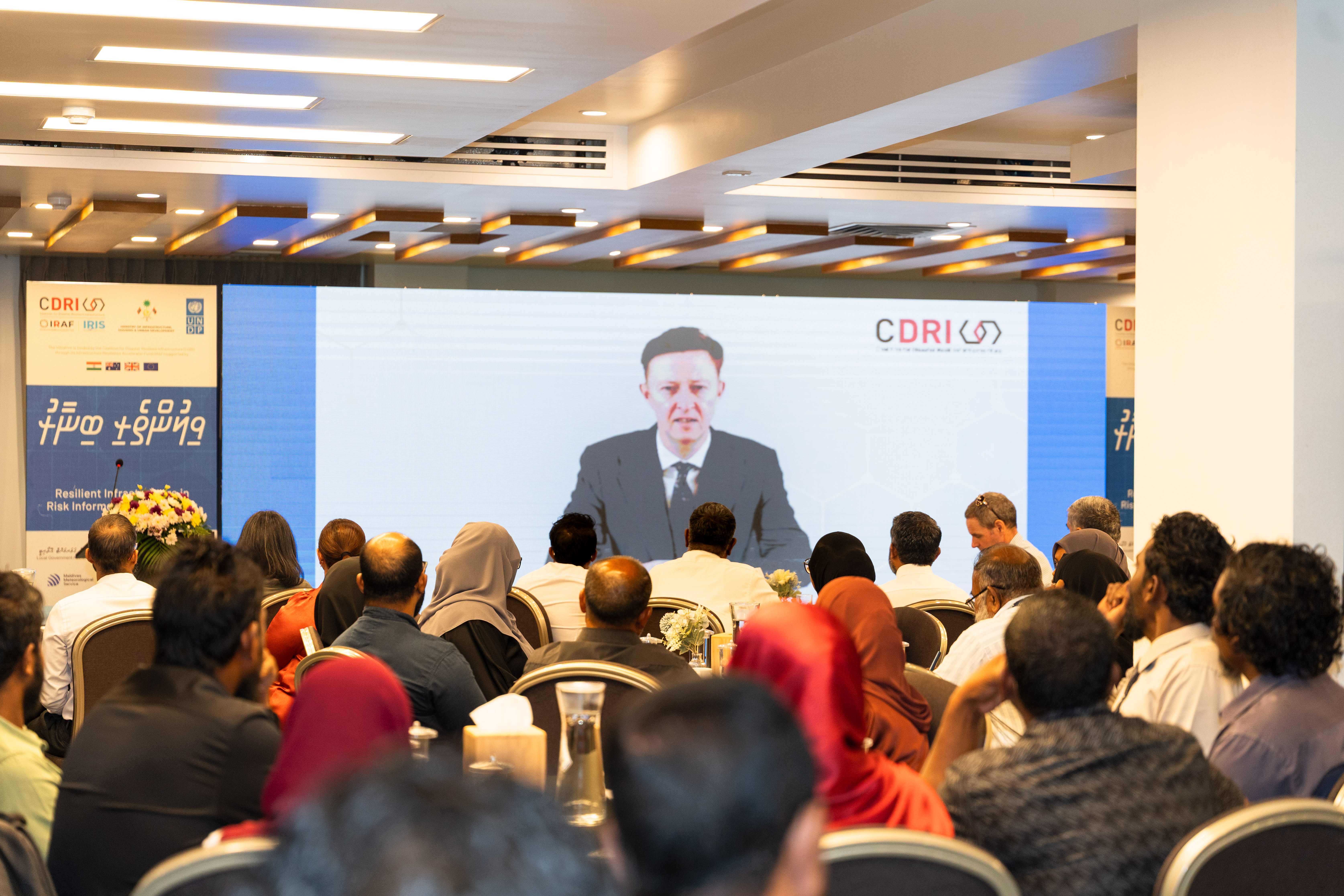 Speaker at a podium addressing a crowd at a conference, with a large screen behind him.