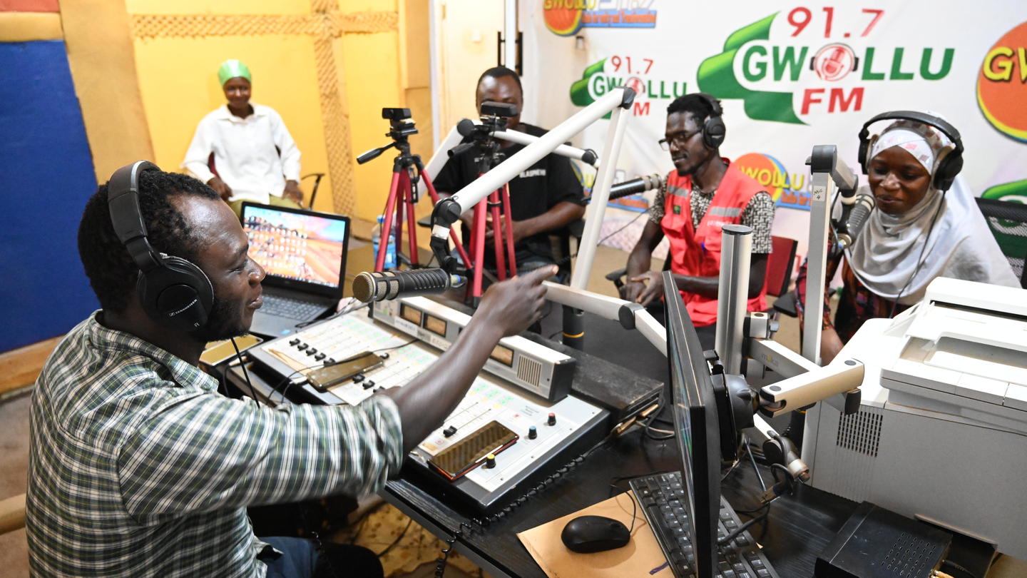 Photograph of a radio studio with hosts around microphones; man in a checkered shirt operates sound mixer.