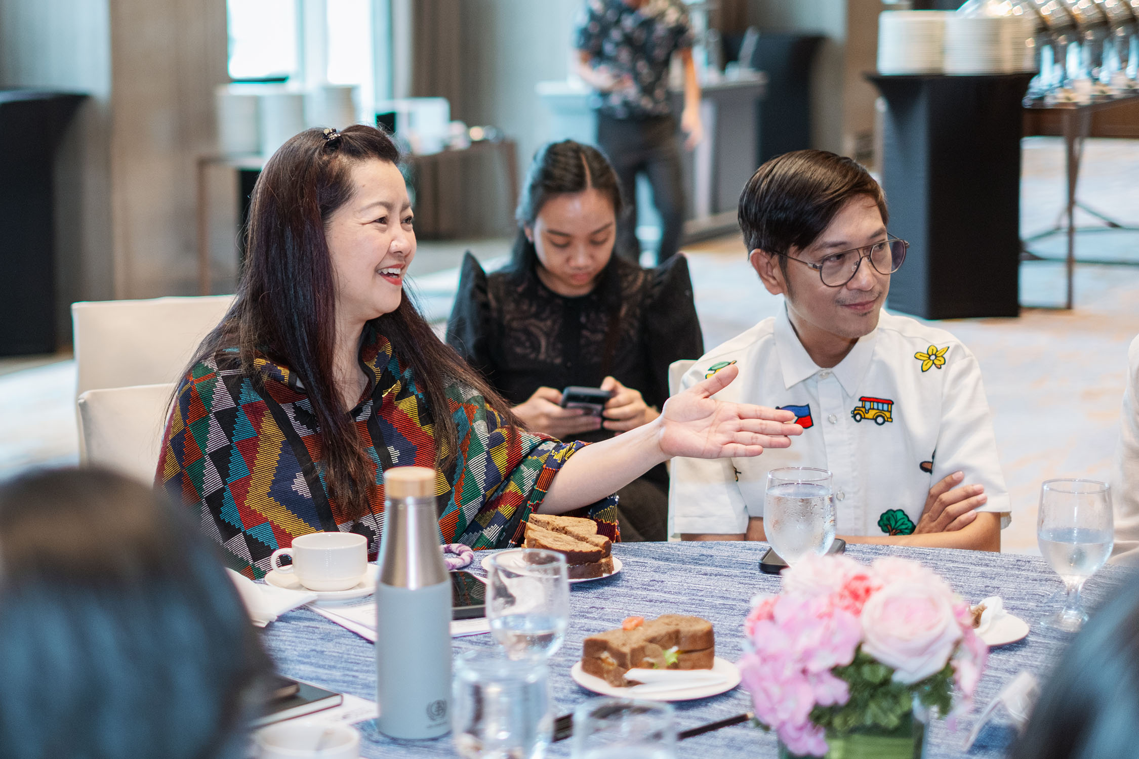 Three people at a bright restaurant table, looking at a document, with pink flowers and desserts.