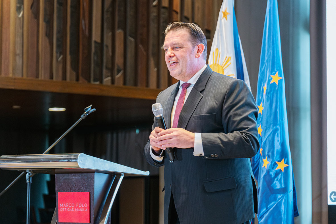 Man in a dark suit speaks into a microphone at a podium, blue EU flags in the background.