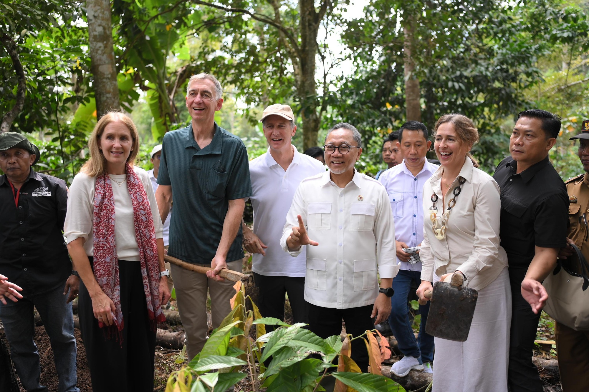 Diverse group of people smiling outdoors in a garden, gathered around a leafy plant.